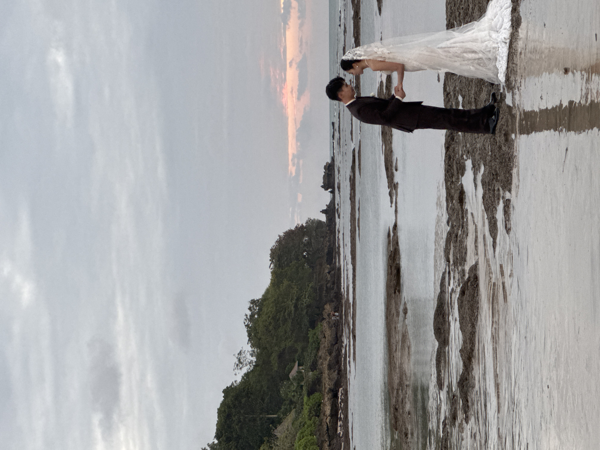 The image depicts a wedding couple on a rocky beach in Kuta Selatan, Indonesia. The couple is positioned centrally, facing each other and holding hands. The groom wears a dark suit and the bride is in a white wedding dress with a veil. The background features a lush, tree-covered hillside to the left, with some small structures visible amongst the trees. The foreground is dominated by shallow water covering a rocky seabed, reflecting the overcast sky. The sky itself is cloudy with a hint of pastel color at the horizon, suggesting either sunrise or sunset. The overall scene is serene and captures a moment of intimacy between the couple amidst a natural coastal environment.