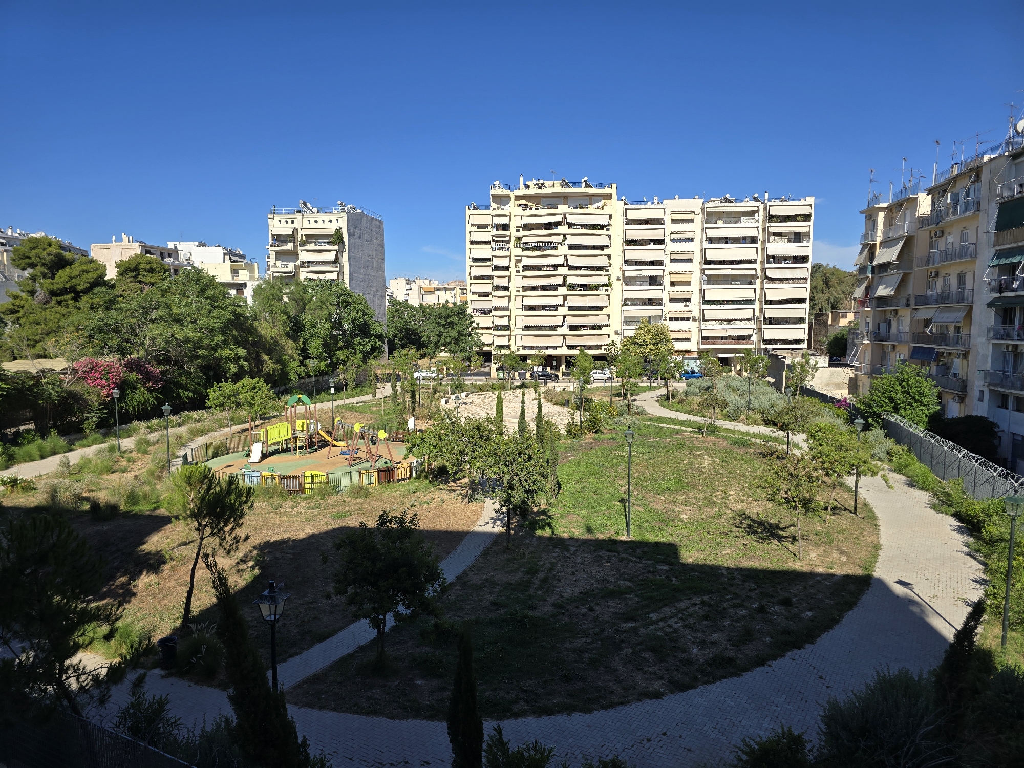 The image depicts a sunny day in an urban park area in Athens, Greece, characterized by modern apartment buildings bordering a green space. In the center of the park, a children's playground with swings, slides, and climbing structures is visible, surrounded by grass and trees. Paved pathways wind through the park, leading to various sections of the green space. Apartment buildings, typical of Athenian architecture with balconies and awnings, rise behind the park, some featuring satellite dishes on their rooftops. The sky is clear and blue, indicating fair weather. The scene is devoid of people, suggesting a quiet moment in the urban landscape. The presence of streetlights, fences, and landscaping indicates a well-maintained public space.