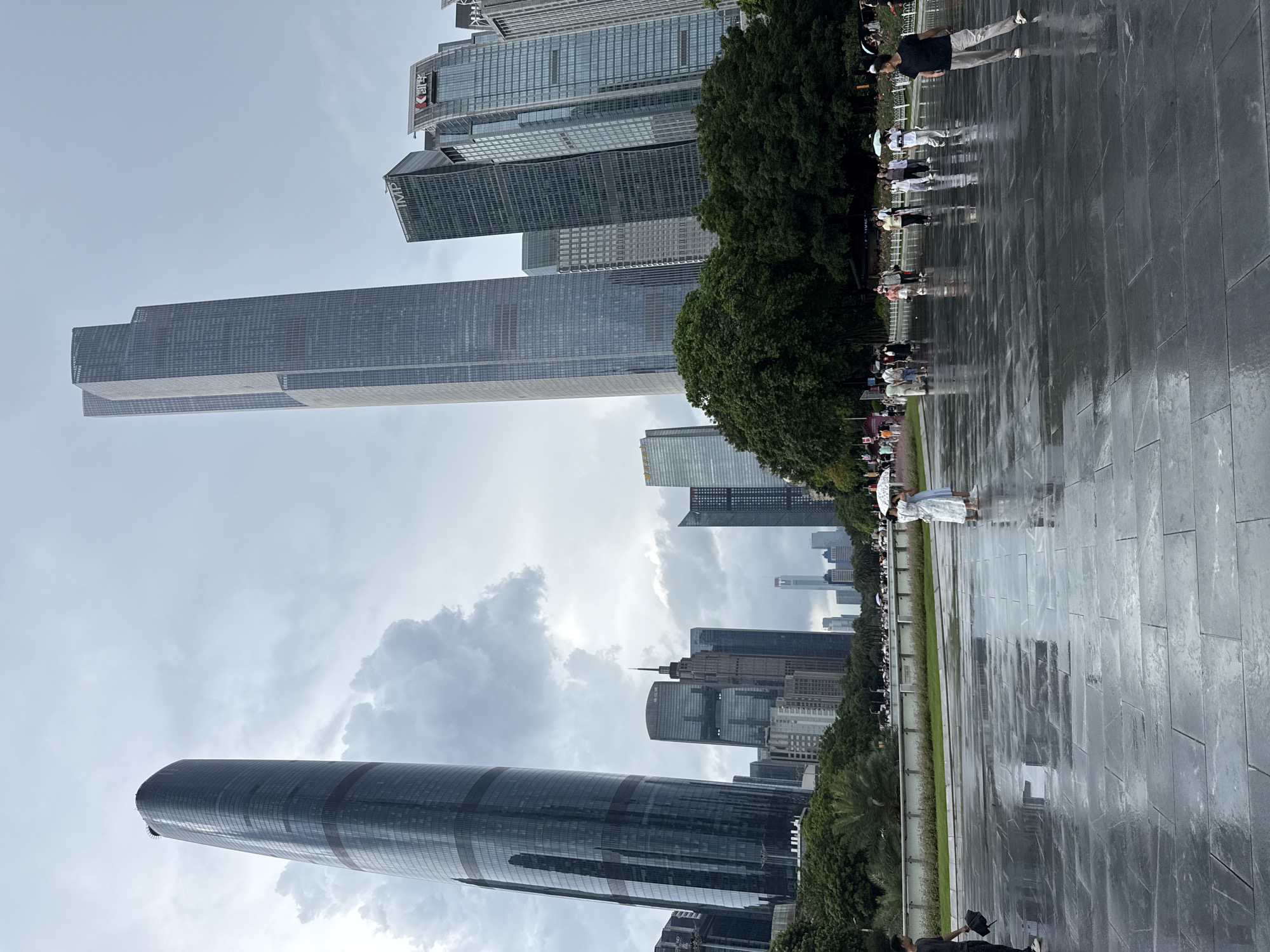 This image captures a wide-angle view of a modern cityscape, likely in the Tianhe District of Guangzhou, China, under an overcast sky. Several prominent skyscrapers dominate the background, characterized by their sleek, glass facades and geometric designs. The foreground features a wet, paved plaza, reflecting the moody sky and the surrounding architecture. A group of people are scattered across the plaza, some holding umbrellas, suggesting recent or ongoing rain.
On the left side of the image, a tall, curved skyscraper with a dark, reflective exterior stands prominently. To its right, a significantly taller, rectangular skyscraper with a grid-like facade pierces the sky, flanked by other contemporary office buildings. Further to the right, the buildings become denser, showcasing a variety of architectural styles. The middle ground is occupied by a lush green park area with mature trees, separating the plaza from the buildings.
In the foreground, the dark, rectangular paving stones of the plaza are visibly damp and reflective, with puddles of water creating mirror-like surfaces. A man in a dark t-shirt and light pants stands on the right, facing away from the camera, his reflection visible in the wet ground. Two women in lighter clothing are walking together on the left side of the plaza, one holding a white umbrella. Other individuals and small groups are visible further back on the plaza, some also with umbrellas. The overall atmosphere is one of urban grandeur combined with the dampness and subdued light of a cloudy day.