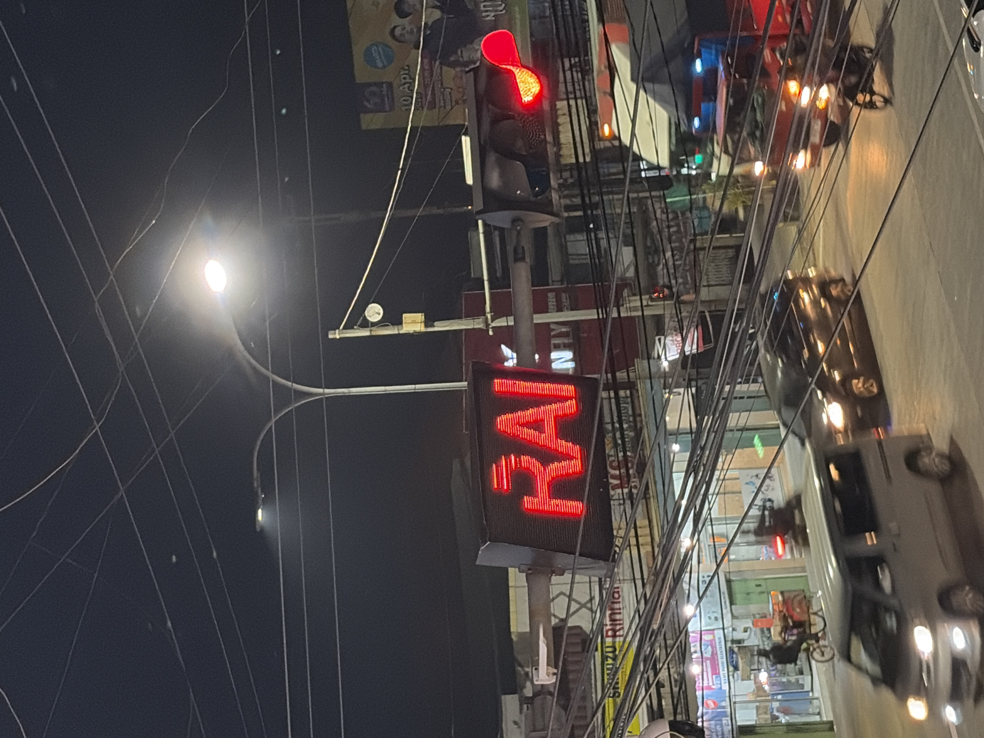 The image captures a nighttime street scene in Batang, Indonesia, with a prominent red LED sign displaying the word "RAI." The sign is mounted on a traffic signal pole, which is adorned with various wires and cables overhead. A traffic light, also red, is visible to the right of the "RAI" sign. The background reveals a street with multiple lanes, where vehicles, including a silver van, a dark car, and a motorcycle, are in motion. Buildings with illuminated storefronts line the street, and a billboard featuring people is also visible in the distance. The overall atmosphere suggests a busy urban environment at night.