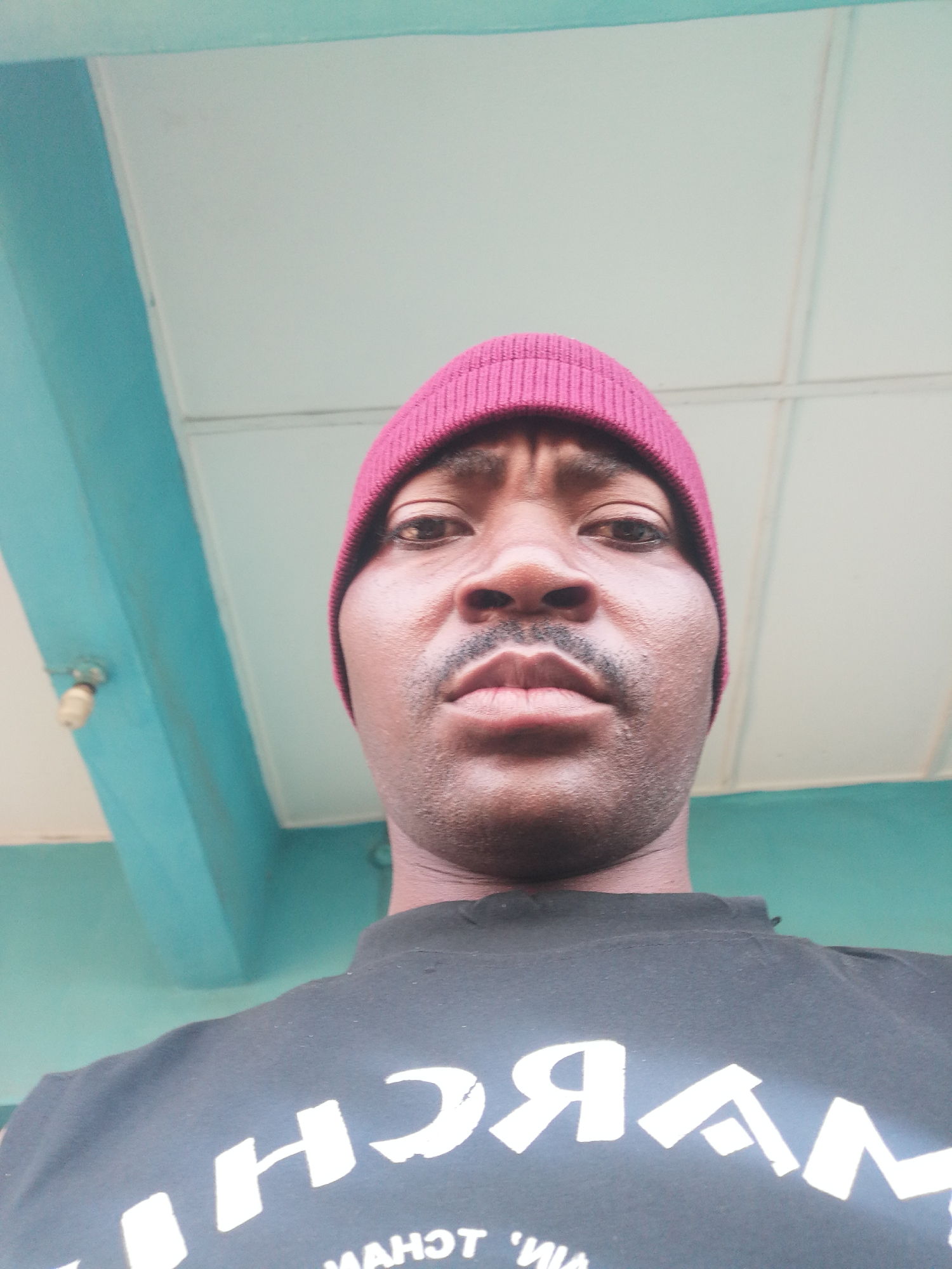 A low-angle, close-up, waist-up selfie of a Black man wearing a maroon beanie and a black t-shirt with white lettering. The man has a mustache. The background consists of a light-colored ceiling and turquoise-colored beams of what appears to be a covered structure. The lighting suggests an indoor or covered outdoor setting. The man's t-shirt has large white lettering that appears to be reversed, but some letters are discernible, such as "H C" and "A N". Below this, there are smaller letters, possibly "M A H O T 'V M A". The scene is consistent with being photographed in a residential or communal area in Ajasse Ipo, Nigeria.