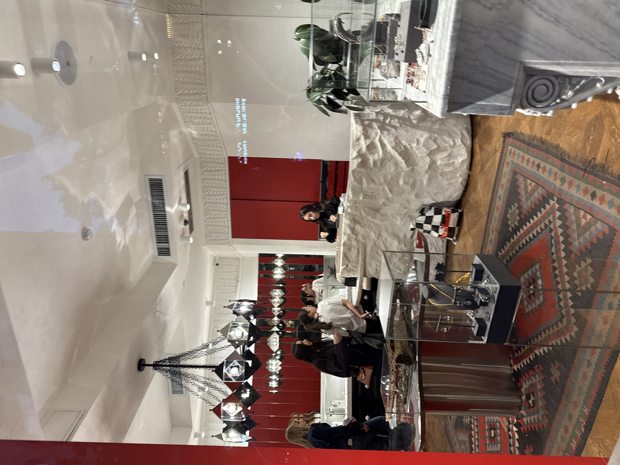The image shows the interior of a retail establishment in Yerevan, Armenia. The interior design features a modern aesthetic with a blend of materials including white walls, polished wood floors, red accent walls, and marble countertops.
Several people are present in the store, seemingly browsing or interacting with staff members. The people appear to be customers or retail staff. The specific activities include looking at merchandise and interacting with other people.
The objects in the store are varied, including jewelry displays, glass display cases with jewelry items, a chandelier, air conditioning vents, and a marble countertop. Additionally, there's a rug with a patterned design on the floor.