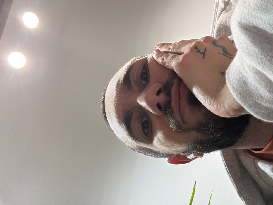 A close-up, low-angle view of a man with dark hair and a beard. His face is angled toward the camera, his left hand resting against his cheek. The skin on his hand has visible tattoos. He wears a grey hoodie, partially visible. In the background, there is a plain white wall and two bright spotlights, slightly out of focus. A couple of green plant stems are visible in the bottom right corner.