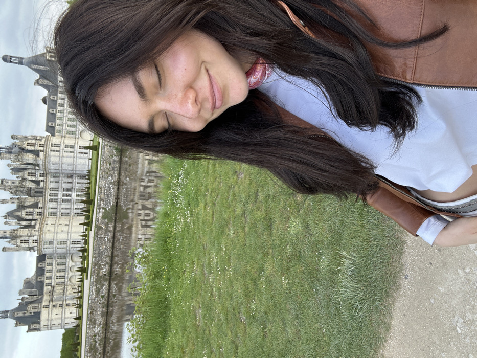 The photo shows a woman with long, dark hair, wearing a brown leather jacket and a white T-shirt with a pink and red bandana around her neck. She is standing in front of the Chateau de Chambord, a grand Renaissance castle in the Loire Valley region of France. The coordinates 47.617, 1.515 indicate the location of the castle. The woman is standing on a path next to a moat. She is looking down at the ground and smiling. Her eyes are closed. The castle is visible in the background. The day is overcast and the water is still. The ground is covered in grass and flowers. The scene is serene and peaceful.