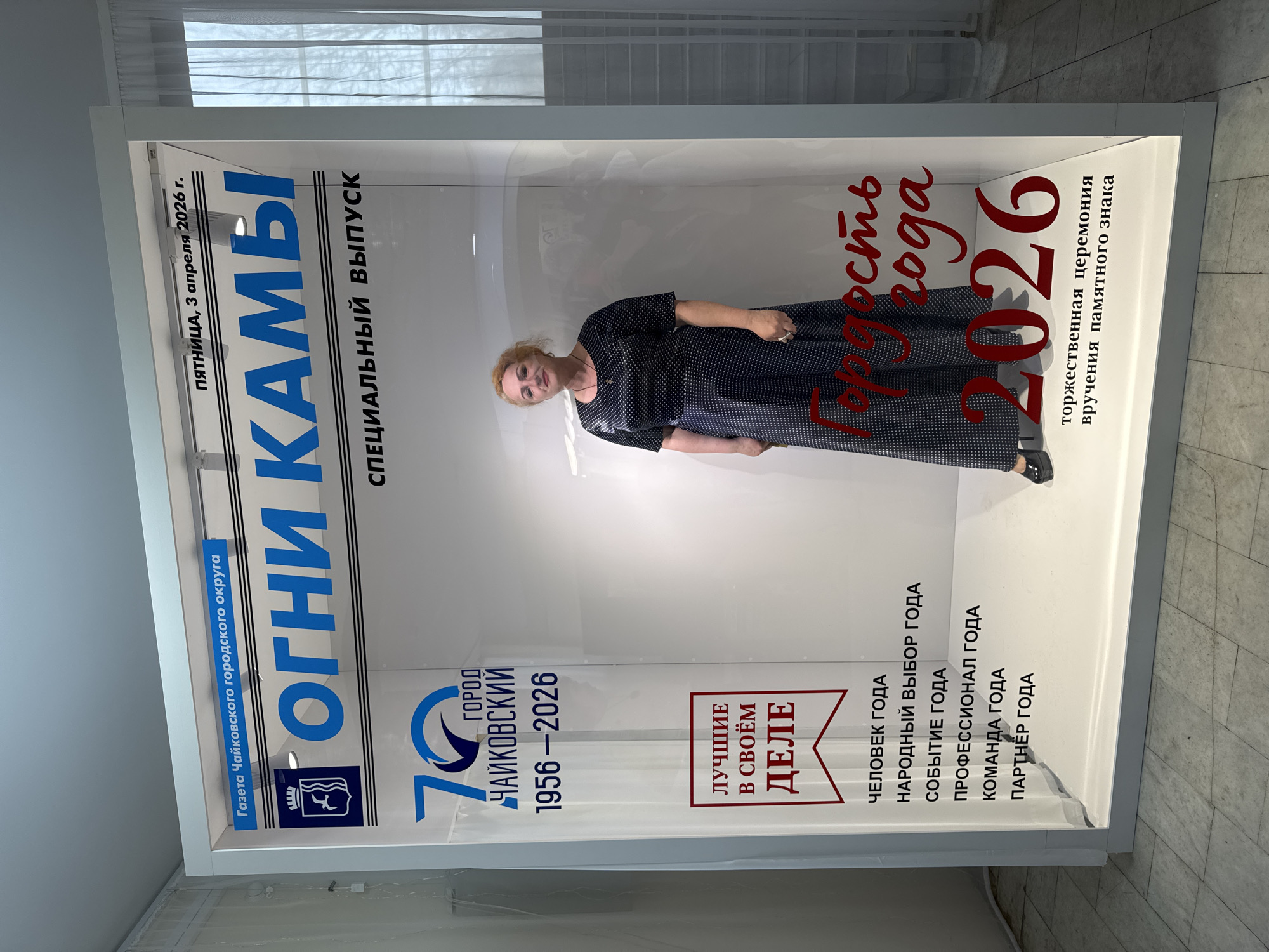 A woman is posing in front of a display case featuring a newspaper. The newspaper is titled "Огни Камь" (Ogni Kamy), which translates to "Lights of Kama". The newspaper is a special issue dated Friday, April 3, 2026, and relates to the city of Chaikovsky, Russia. The display celebrates the 70th anniversary of the city (1956-2026).
The newspaper also lists categories like "Man of the Year", "People's Choice of the Year", "Event of the Year", "Professional of the Year", "Team of the Year", and "Partner of the Year". A red inscription reads "City of the Year" along with the year "2026". The display also advertises a ceremony for the presentation of a commemorative sign. The location is within the city of Chaikovsky, Russia.