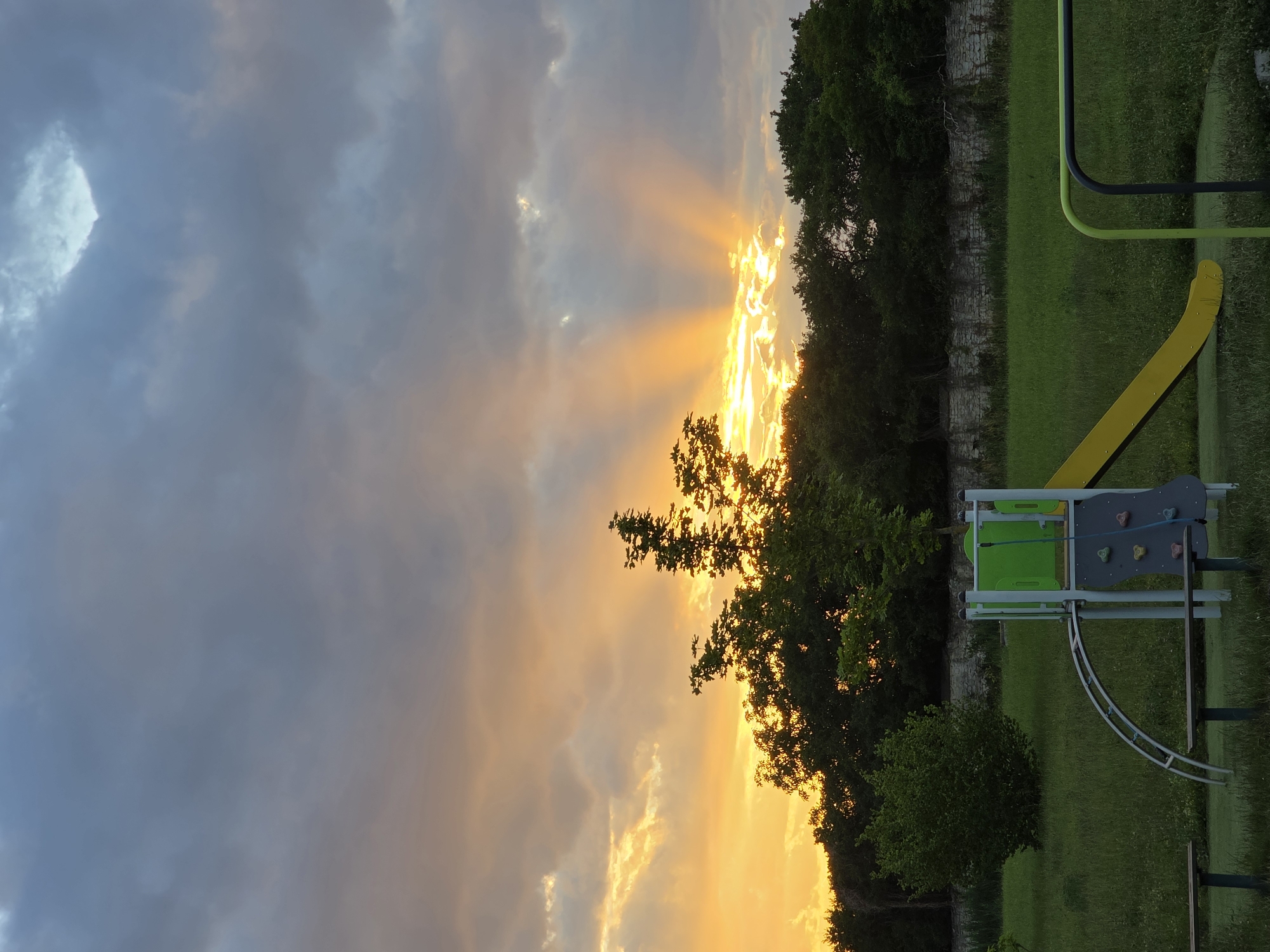 This image depicts a playground scene at dusk, with a dramatic sunset casting golden rays through the clouds. In the foreground, a yellow slide with a green climbing wall and a silver archway is partially visible. Two dark benches are situated in front of the playground equipment. The ground is covered in green grass, with a textured, dark green surface beneath the slide. A low stone wall runs horizontally across the middle ground, behind which is a dense line of dark green trees. The sky is filled with a mix of dark gray and light blue clouds, with bright yellow and orange hues emanating from the sunset. The overall mood is serene and somewhat dramatic due to the celestial display. There are no people present in the image.