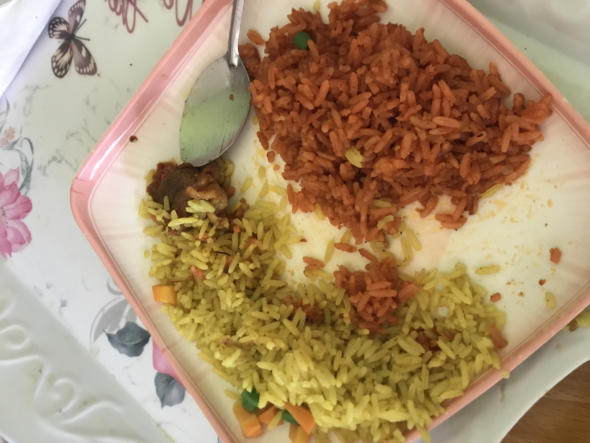 An overhead, close-up photograph depicts a meal served on a rectangular, off-white plate with a light pink rim. The plate is positioned on a decorative tray featuring white embossed scroll patterns, pink floral motifs, grey leaves, and a brown butterfly illustration. The meal consists of two distinct types of rice. On the left, a mound of yellowish-green rice is visible, containing diced orange carrots and green peas. On the right, a separate mound of orange-red rice, possibly jollof, is present, with one green pea visible. Between these two rice portions, towards the top-center, is a small quantity of stewed meat in a reddish-brown sauce. A silver-colored metal spoon rests on the plate, partially overlapping the orange-red rice. The surrounding surface is brightly lit, suggesting an indoor setting. This scene is located in the city of Jos, Nigeria.