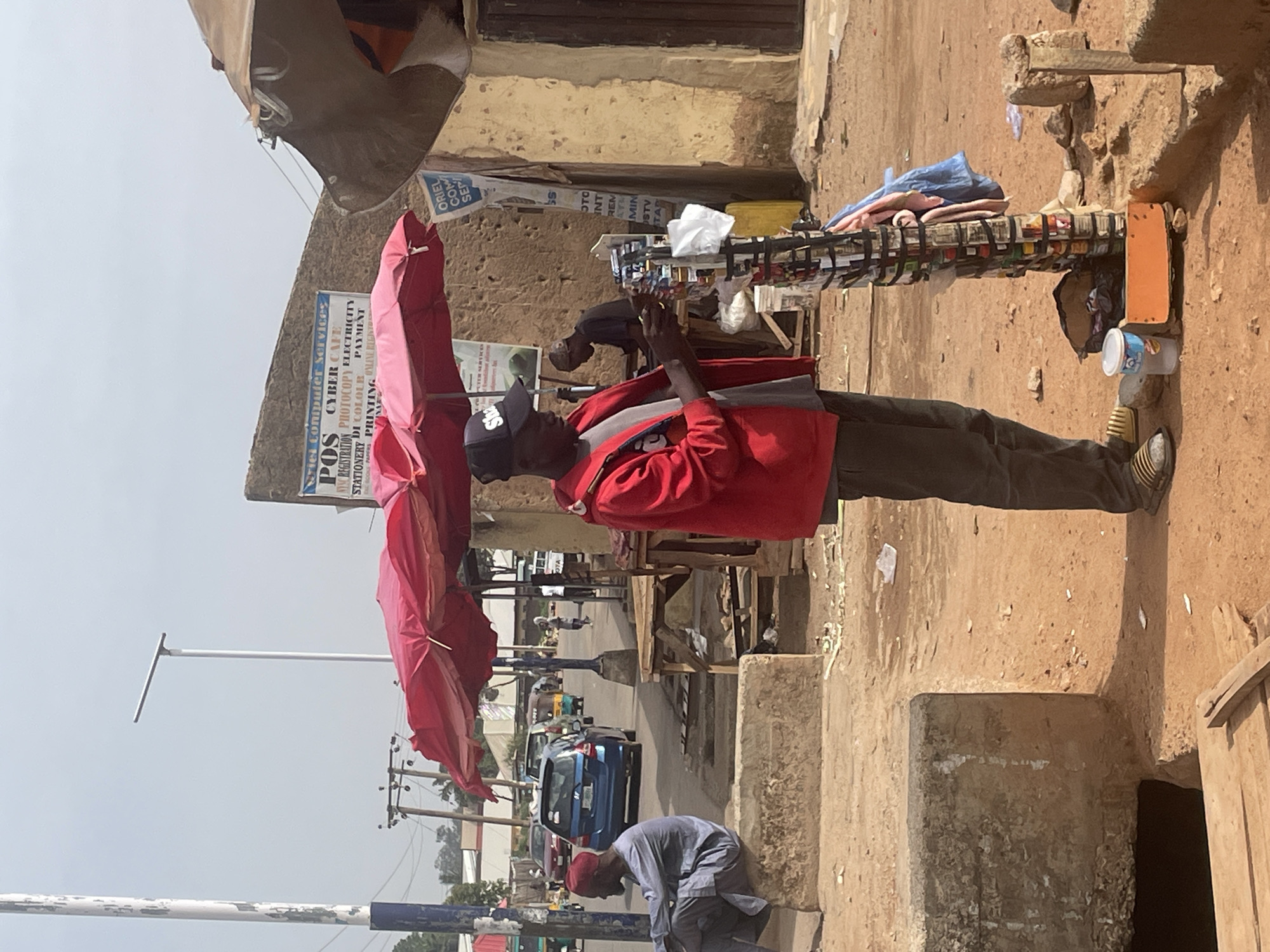 The image depicts a street scene in Jos, Nigeria. A man in a red jacket and cap stands next to a display of small goods, possibly for sale, sheltered by a red umbrella. He appears to be using a mobile phone. Behind him, another man is bent over, possibly tending to the display.
The backdrop shows a building with faded signage advertising "Cyber Cafe" and "Computer Services," offering services like photocopying, printing, and payment options. The street features passing vehicles, including a blue car and a "keke". A light pole is visible on the left side of the frame. On the right side, there is another building and small cans.
The overall environment suggests a bustling street market or commercial area in a developing urban setting.