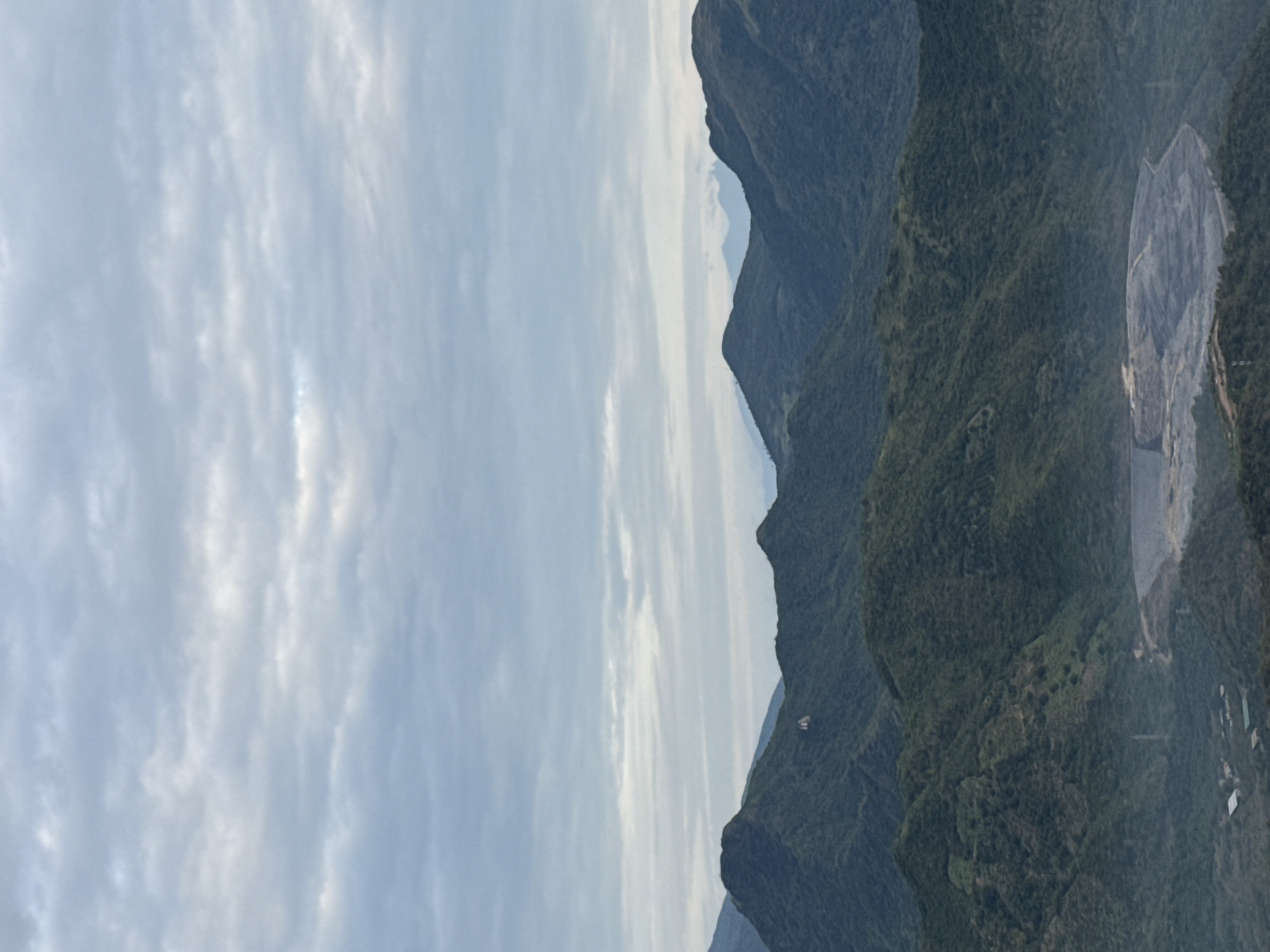 The image shows a view of mountains under a cloudy sky, likely taken near Bac Nha Trang Ward, Vietnam. The mountains are covered in dense green vegetation. A large, disturbed area of lighter-colored terrain, potentially a quarry or construction site, is visible on one of the slopes. The sky is predominantly gray with patterns of clouds.
