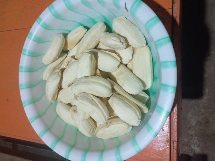 A white plastic bowl, adorned with wavy green stripes, is filled with numerous peeled, small, and light-colored fruits. The fruits are elongated and have a creamy white to pale yellow hue, resembling prepared bananas or plantains. They are piled loosely within the bowl, showcasing their smooth texture.
The bowl is situated on a surface that appears to be an orange-brown wooden table or floor, visible predominantly to the left of the bowl. To the right, the background is darker and less distinct, possibly indicating a shadow or another environmental element. The lighting suggests an indoor setting. There are no people visible, and no specific activity is taking place other than the presentation of the prepared fruit. No text is visible in the image. The provided location context for this scene is Pubian, Indonesia.