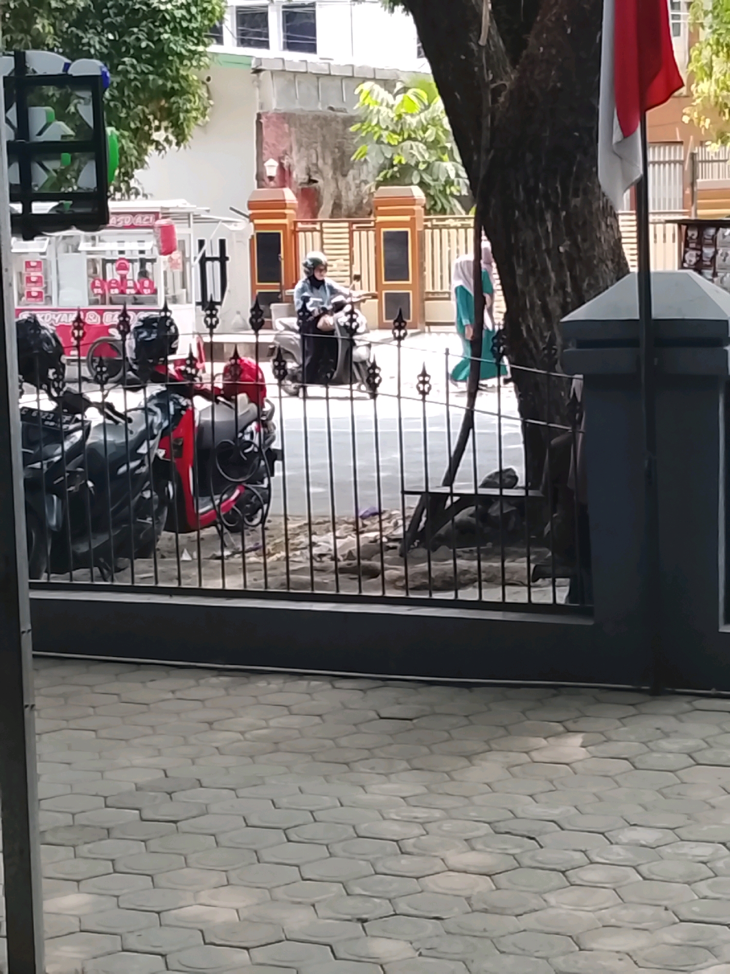 The image depicts a street scene in Slawi, Indonesia, viewed through a black metal gate. In the foreground, the gate's horizontal bars are prominent, with a hexagonal paver walkway visible beyond. Behind the gate, several motorcycles are parked, some with helmets on their seats. A food cart, possibly selling ice cream, is visible to the left, with red and white branding. Further back, a person wearing a helmet rides a scooter, and another individual in a green hijab walks past a tree with a rough bark. A building with white walls and some red accents is in the background, along with a portion of the Indonesian flag. The lighting suggests it is daytime.
