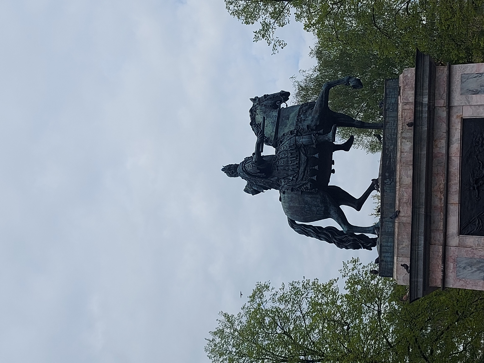 The image shows an equestrian statue in what is likely Saint Petersburg, Russia. The statue depicts a rider on a horse, both elaborately detailed. The statue rests on a marble base. The statue is surrounded by trees. The sky is overcast. A bird is visible in flight below the statue. The view is from below, looking up at the statue.
