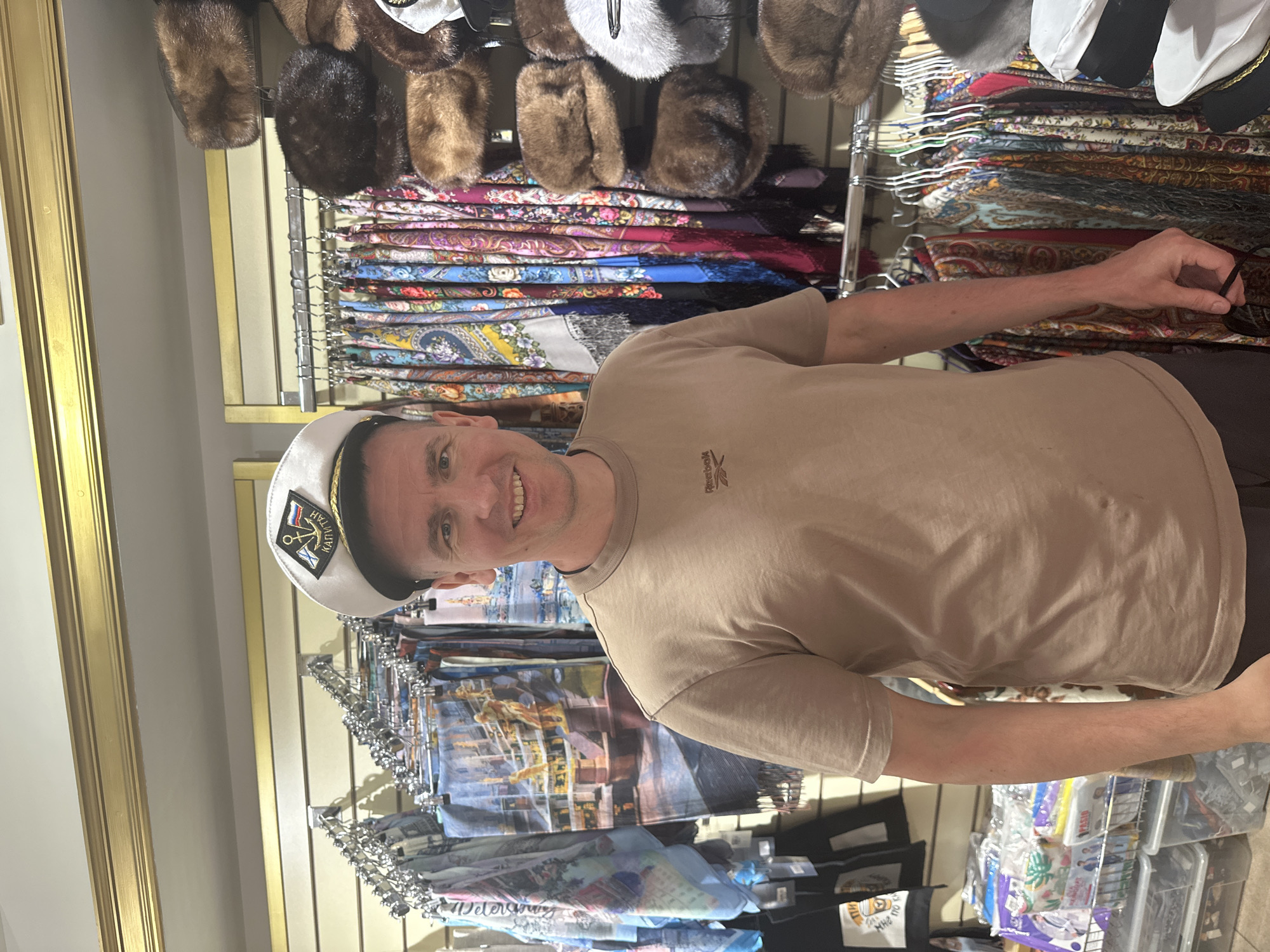 A middle-aged Caucasian man is standing inside a shop in Saint Petersburg, Russia. He is wearing a beige t-shirt and a white sailor's cap with the word "КАПИТАН" (Captain) embroidered on it. He is smiling and looking at the camera.
Behind him, there is a display of fur hats on the top row and several rows of colorful shawls and scarves. Some items have "Petersburg" printed on them. There are other products in the lower right part of the image. The store appears to be a souvenir shop.