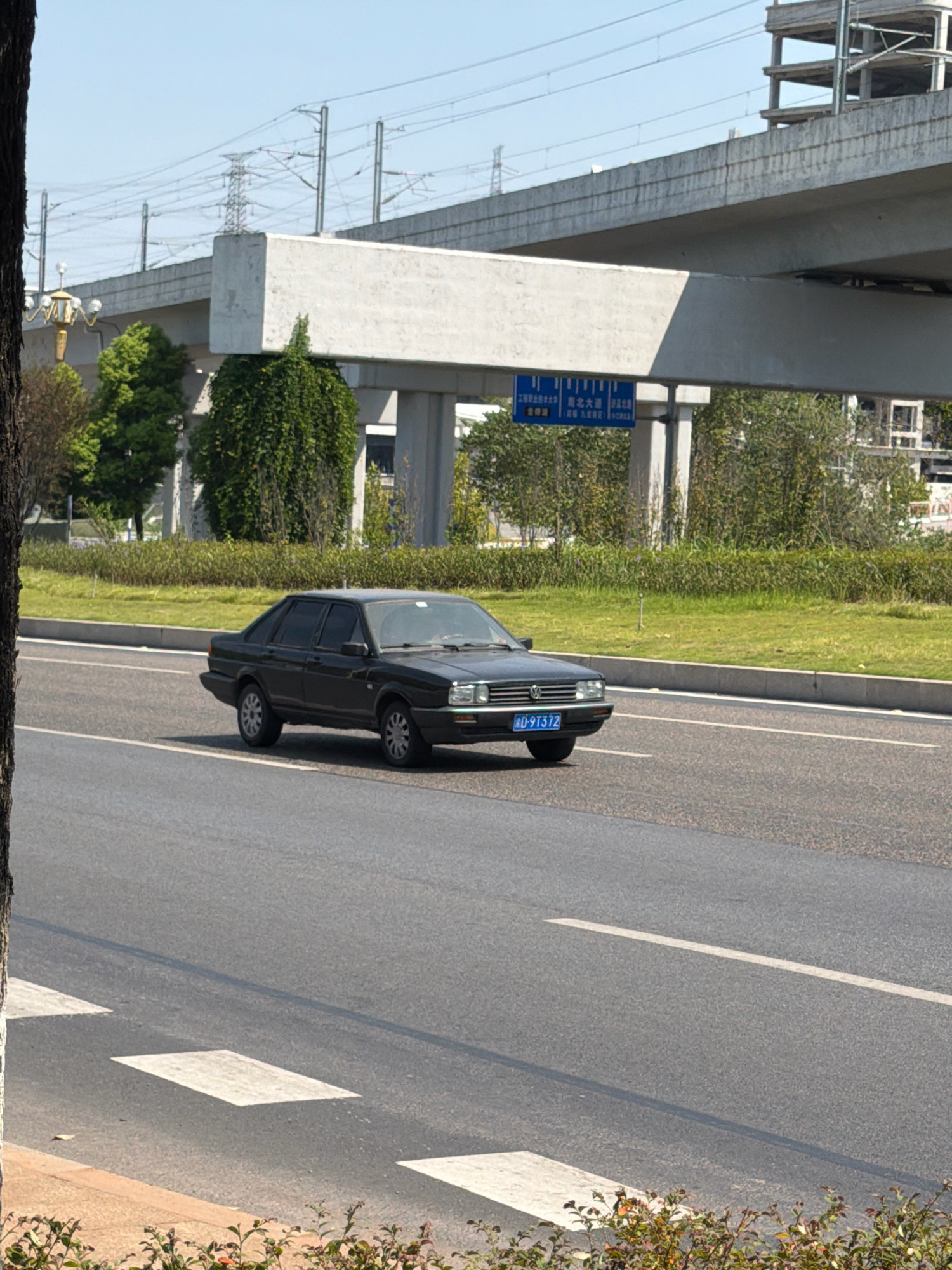 A dark colored sedan is driving on a paved road under an elevated highway structure. The car has a blue license plate with white characters. The road has white lane markings and a crosswalk. The environment includes green grass, trees, and a construction site in the background. Overhead, power lines and electrical towers are visible. A blue sign with white text is mounted on a pole under the highway. The scene appears to be in Jiangjin, China.