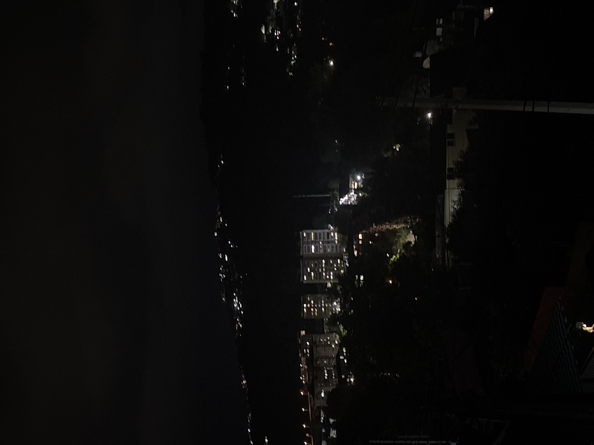 The image depicts a nighttime scene in Sochi, Russia. It appears to be a view from a high vantage point, overlooking a body of water and buildings illuminated by lights. The overall impression is one of urban tranquility at night.
The primary subjects are the buildings with their lit windows, suggesting residential or commercial spaces. The dark water to the left likely reflects the lights, creating shimmering streaks that add a dynamic element to the scene. Trees and foliage are also visible, breaking up the cityscape and adding a touch of nature to the urban environment. A few scattered lights in the distance could indicate further development or distant areas.
The time of day is clearly night, given the darkness and the illuminated windows. There are no discernible weather conditions mentioned or visible, but the overall atmosphere suggests a clear night. Location cues are limited to the fact that the scene is in Sochi, Russia, and the visual elements are consistent with an urban coastal environment at night.
There is no discernible activity or event taking place beyond the general illumination of a city at night. No people are clearly visible, and the focus is on the static elements of the landscape and architecture.
Notable details include the long streaks of light on the water, the pattern of lit windows in the multi-story buildings, and the contrast between the dark night sky and the illuminated structures. The image is predominantly dark, with the lights serving as points of interest.
There is no visible text in the image.