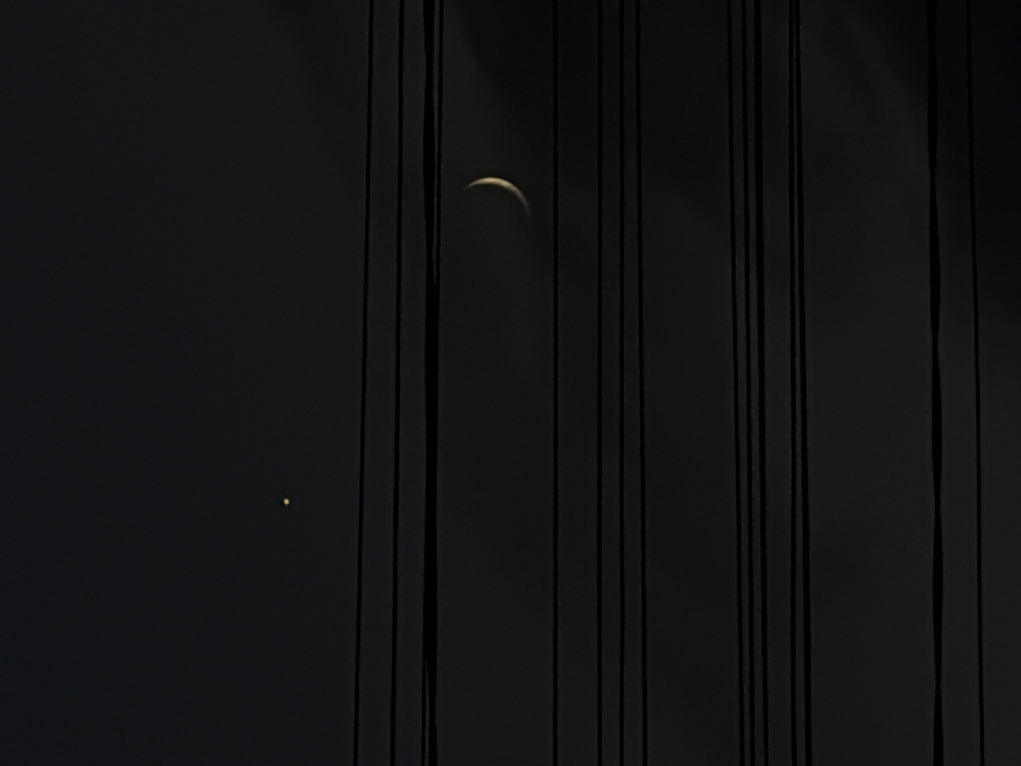 The image shows a dark sky with several horizontal lines that appear to be power lines. In the upper left portion of the sky, a small, bright yellow point of light is visible, likely a planet or star. In the middle right of the sky, a crescent moon with a faint glow is also visible. The overall scene is one of a night sky with celestial bodies and man-made infrastructure. No people or specific landmarks are visible. The location in Dongaron, Russia cannot be determined from the image alone.