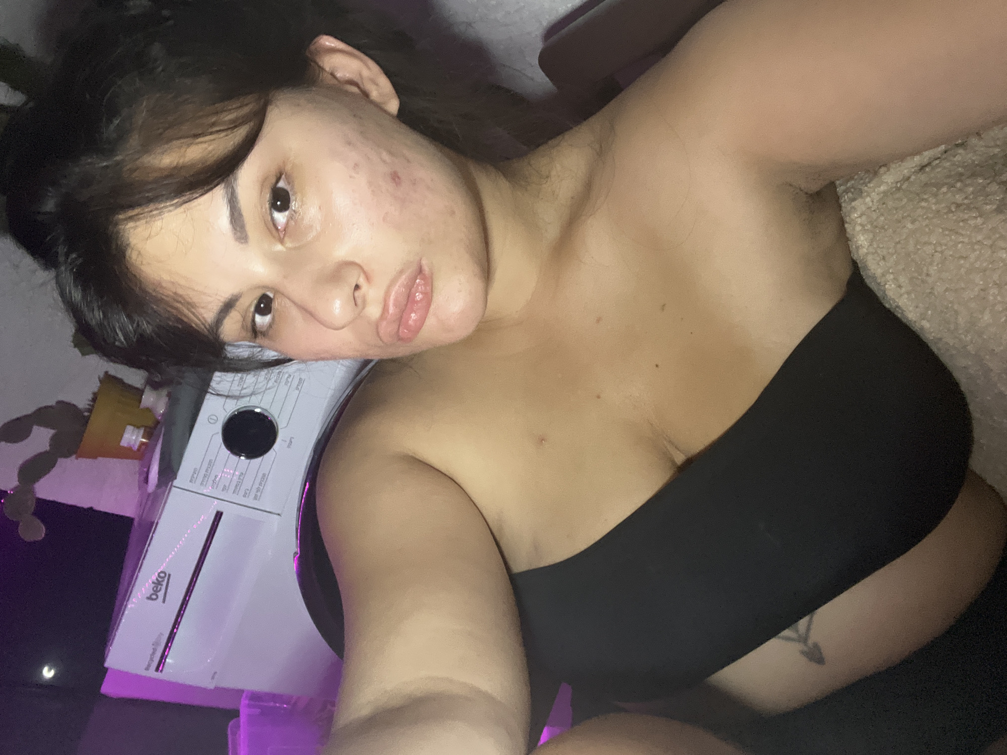 The image displays a close-up selfie taken indoors, presumably in an apartment in Kiryat Bialik, Israel. The subject is a young woman with dark hair pulled back, positioned centrally in the frame. She is wearing a black strapless top. Her face is turned slightly towards the camera, and she appears to be looking directly at it. Visible on her face are several blemishes and acne scars.
In the background, a white Beko washing machine is partially visible, illuminated by purple ambient lighting. A cactus in a yellow pot sits to the upper right of the washing machine. The overall environment appears to be a domestic setting, possibly a laundry area or bedroom, given the presence of household appliances and soft furnishings. The lighting is dim, with the purple glow from a light source creating a distinct mood. The image is taken from a slightly elevated angle, looking down at the subject.