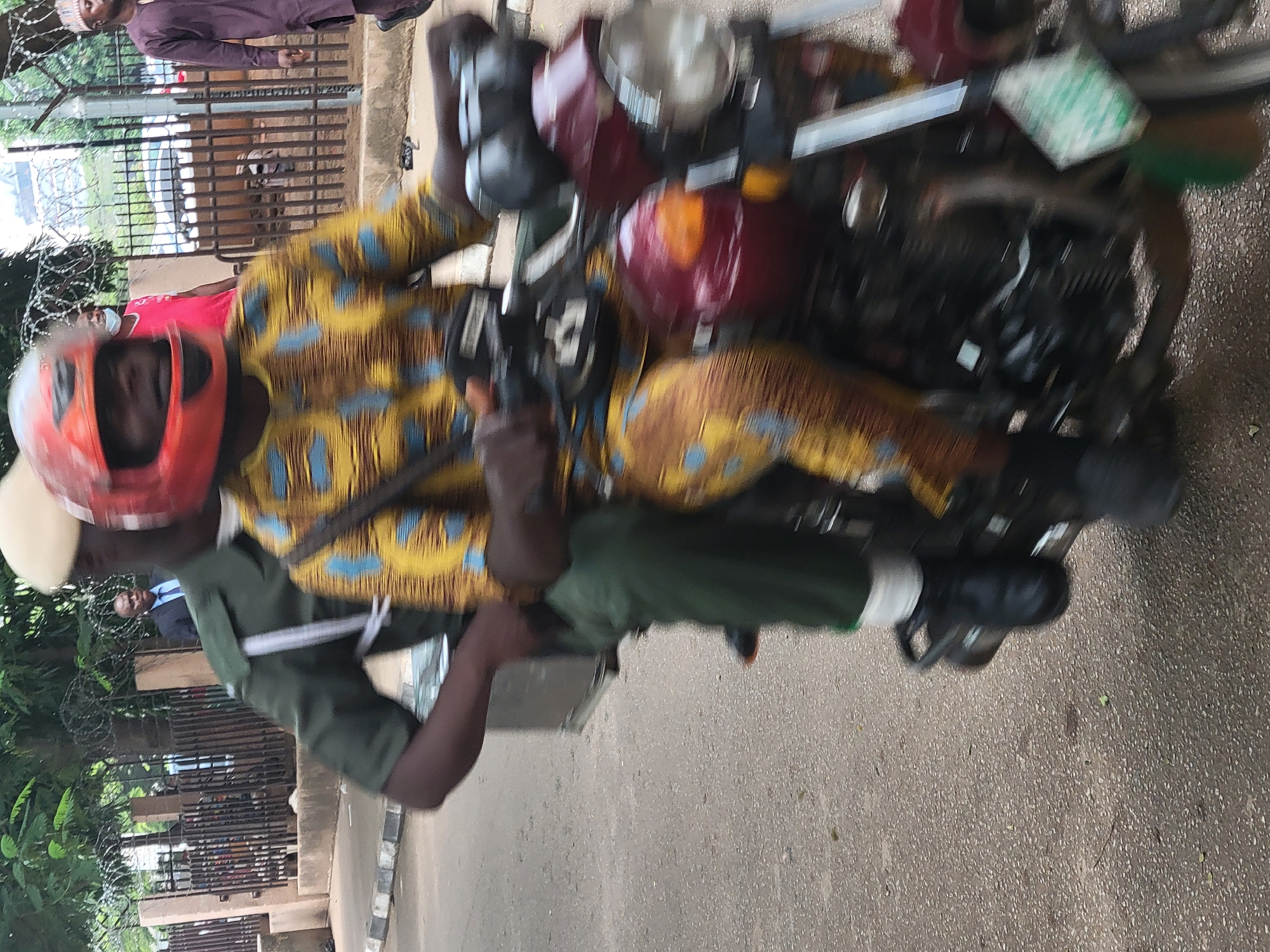 A dynamic, motion-blurred outdoor shot captures two men riding a motorcycle. The image is oriented vertically, showing the subjects and vehicle from a slightly elevated side perspective, facing left.
The man driving the motorcycle wears a vibrant, patterned yellow and blue long-sleeved shirt and matching trousers, along with a bright orange helmet that obscures most of his face, revealing only his chin and mouth. His hands are on the handlebars. Seated behind him, the passenger wears a dark green uniform, consisting of a shirt and trousers, and a light-colored cap. He has a strap or bag across his chest. His posture suggests he is holding onto the driver.
The motorcycle itself appears to be a standard model, predominantly dark red or maroon in color, with a chrome headlight and other metallic components. A green, rectangular object, possibly a license plate or permit, is visible near the front of the motorcycle, though any text on it is illegible due to blur.
The background features a dark metal fence topped with barbed wire, suggesting a secure perimeter. Beyond the fence, a few indistinct figures can be seen, including a man in a purple traditional robe and cap on the far left, and another man in a suit jacket and tie further back. Green foliage and trees are visible behind the fence and in the distance. The ground beneath the motorcycle is paved asphalt or concrete.
The significant motion blur across the entire image indicates that the motorcycle was moving at the moment the photo was taken, conveying a sense of speed and action. The scene appears to take place during daylight hours under clear weather conditions, consistent with the location context of Abuja, Nigeria.