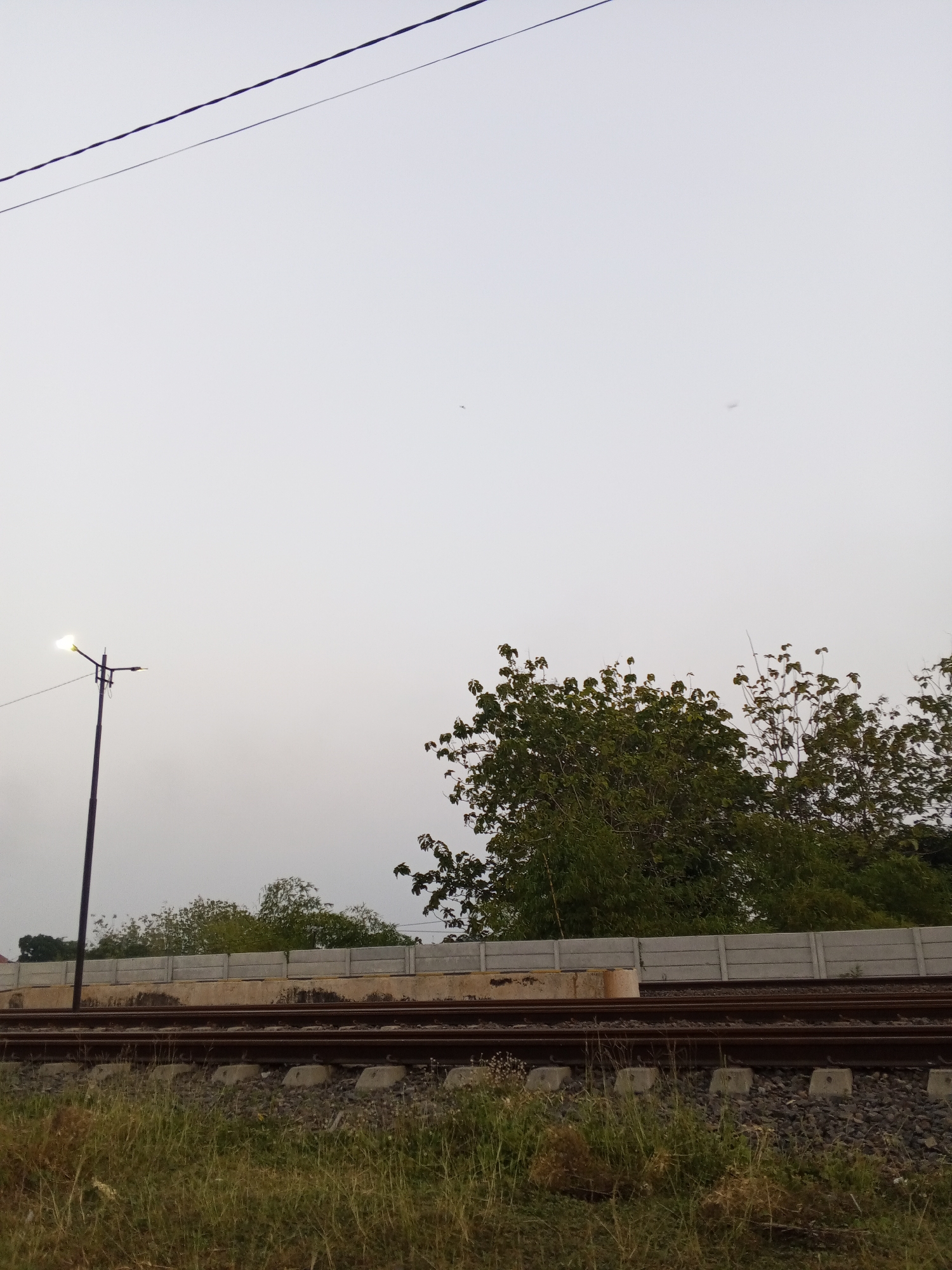 The image shows a view of railroad tracks in Pakusamben, Indonesia, under a pale, overcast sky. A single dark lamppost with its light on stands to the left of the frame. Beyond the tracks, a concrete wall runs horizontally, with dense green trees and foliage behind it. The foreground consists of overgrown grass and weeds. In the upper portion of the image, two dark power lines are visible against the sky. The scene appears to be a rural or semi-urban area, likely near a train line, with no people present.