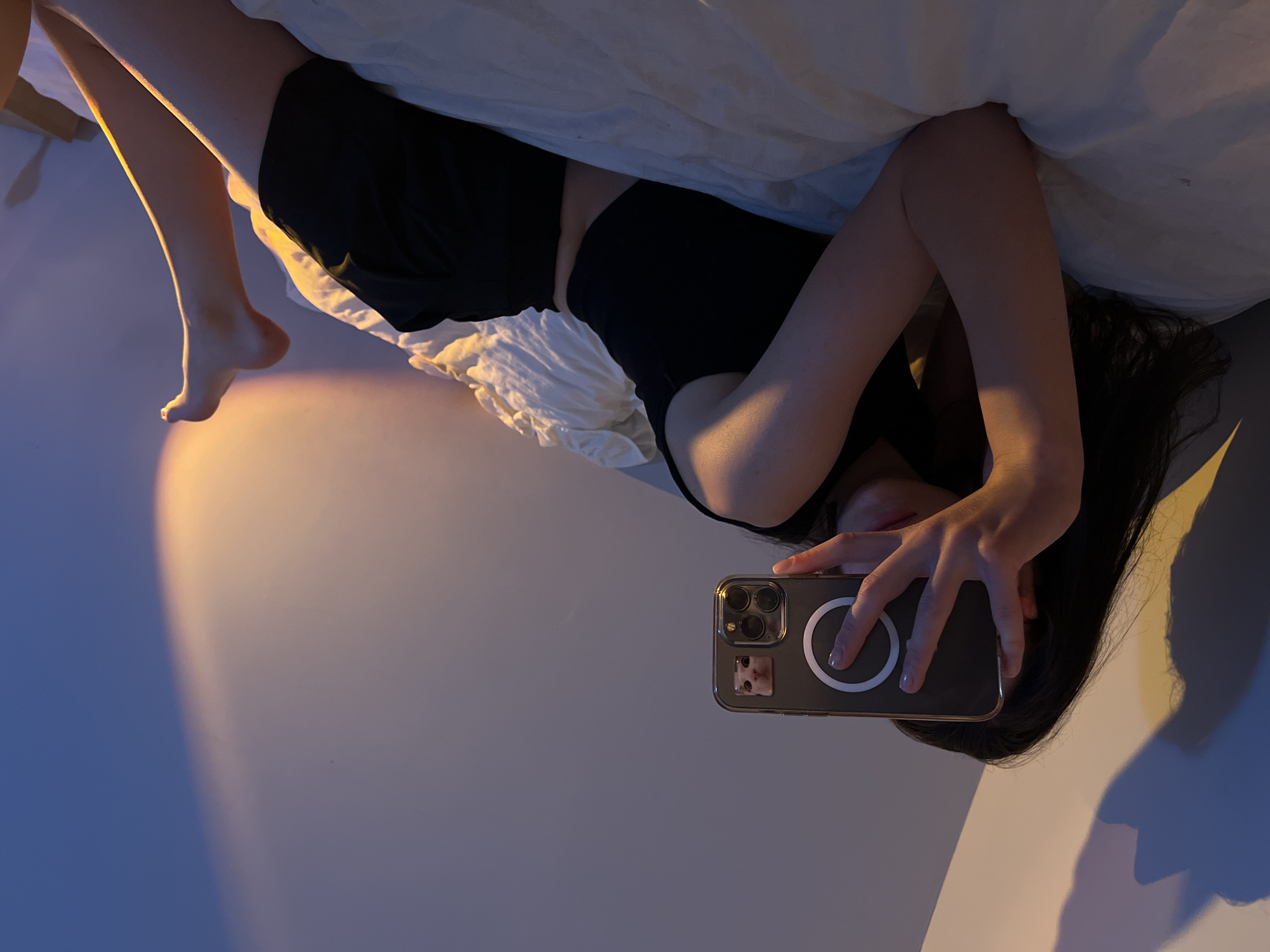 A person is lying on a bed, taking a selfie with a smartphone. The person is wearing a black tank top and black shorts. The phone is held horizontally, and the person's face is partially obscured by their hand. The background is a wall with a shadow cast upon it, illuminated by warm lighting. The bed appears to be covered with white sheets. The scene is indoors, likely a bedroom. The image does not contain any recognizable landmarks or features that would indicate its location in Ternopil, Ukraine.