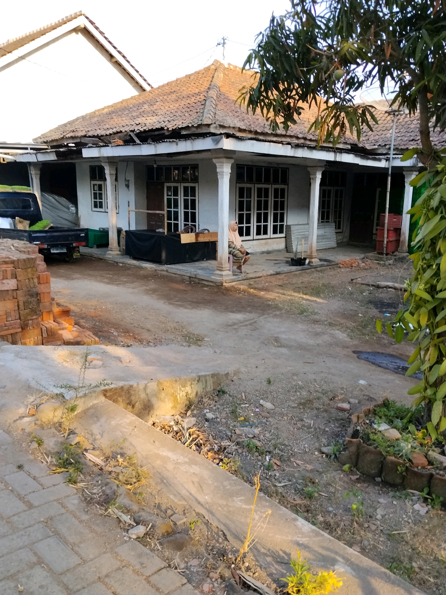 The image captures a front yard scene in Tlogosari, Indonesia, with a focus on a modest house. The house features a tiled roof with some visible wear and tear, and a covered porch supported by white pillars. Several windows with white frames are visible on the house's facade.
In the foreground, a paved walkway runs along the bottom left of the image, transitioning to a dirt and gravel yard. To the left, a stack of orange bricks is piled high, suggesting ongoing construction or renovation. A dark-colored pickup truck with a black tarp covering its bed is parked near the house.
A woman wearing a headscarf and a patterned top sits on a pink chair on the porch, facing towards the left of the frame. She appears to be looking away from the camera.
The yard is somewhat unkempt, with patches of dry grass, scattered debris, and some potted plants. A large tree with dark green leaves dominates the right side of the image, partially obscuring the house and its surroundings. The overall impression is a residential area, likely in a tropical climate given the vegetation. The lighting suggests it is either early morning or late afternoon, with a warm, diffused sunlight.