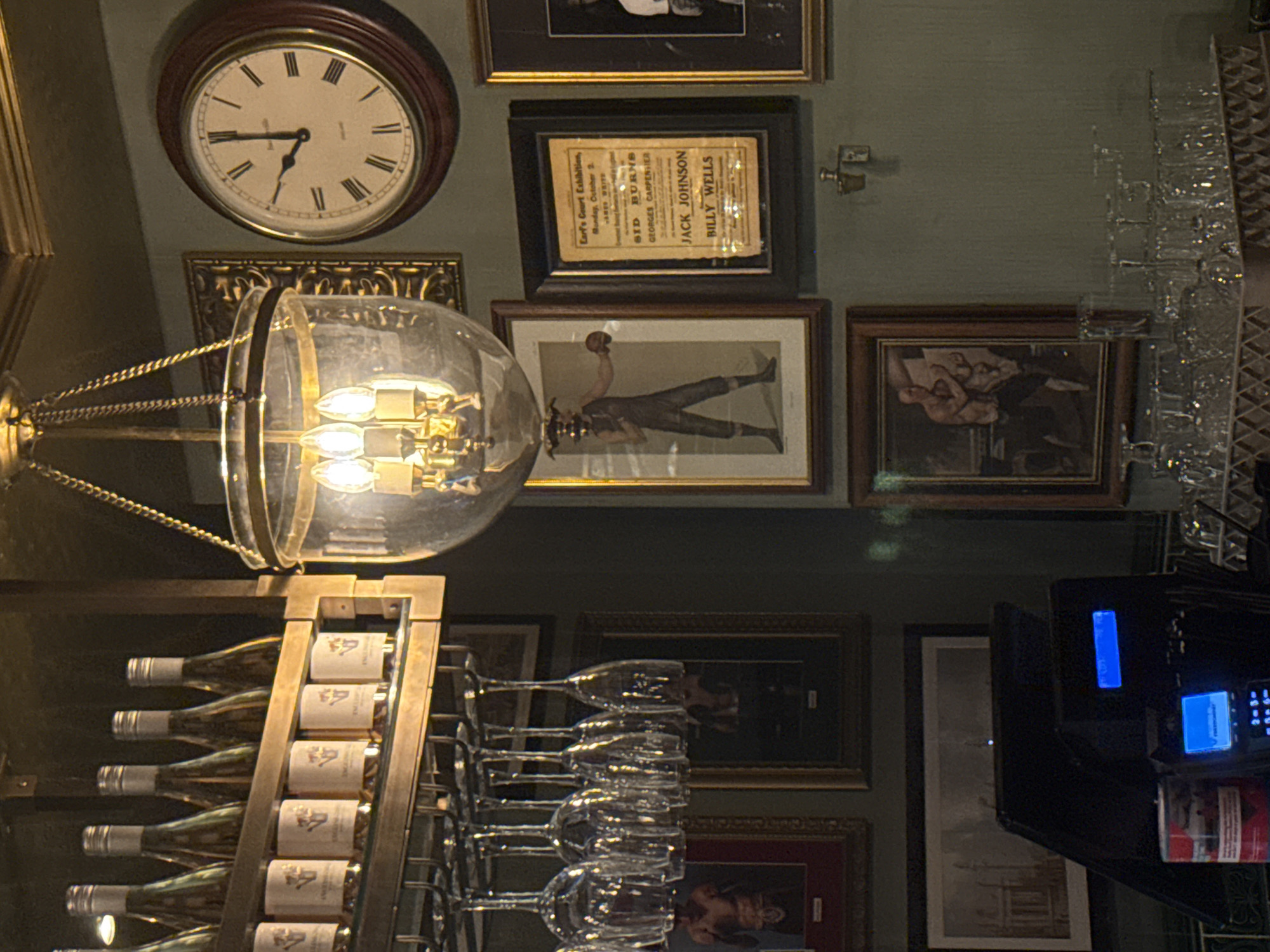 The image depicts the interior of a bar or restaurant in London, United Kingdom. A wine rack with bottles of wine and stemmed glasses dominates the lower left. Above, a vintage clock shows approximately 7:48. Framed images are displayed on the wall, including an advertisement for an "Earl's Court Exhibition" featuring names like "Sid Burns", "Georges Carpentier", "Jack Johnson", and "Billy Wells". Other framed images include portraits and what appear to be boxing scenes. A cash register is visible in the lower right corner, indicating the location is a business.