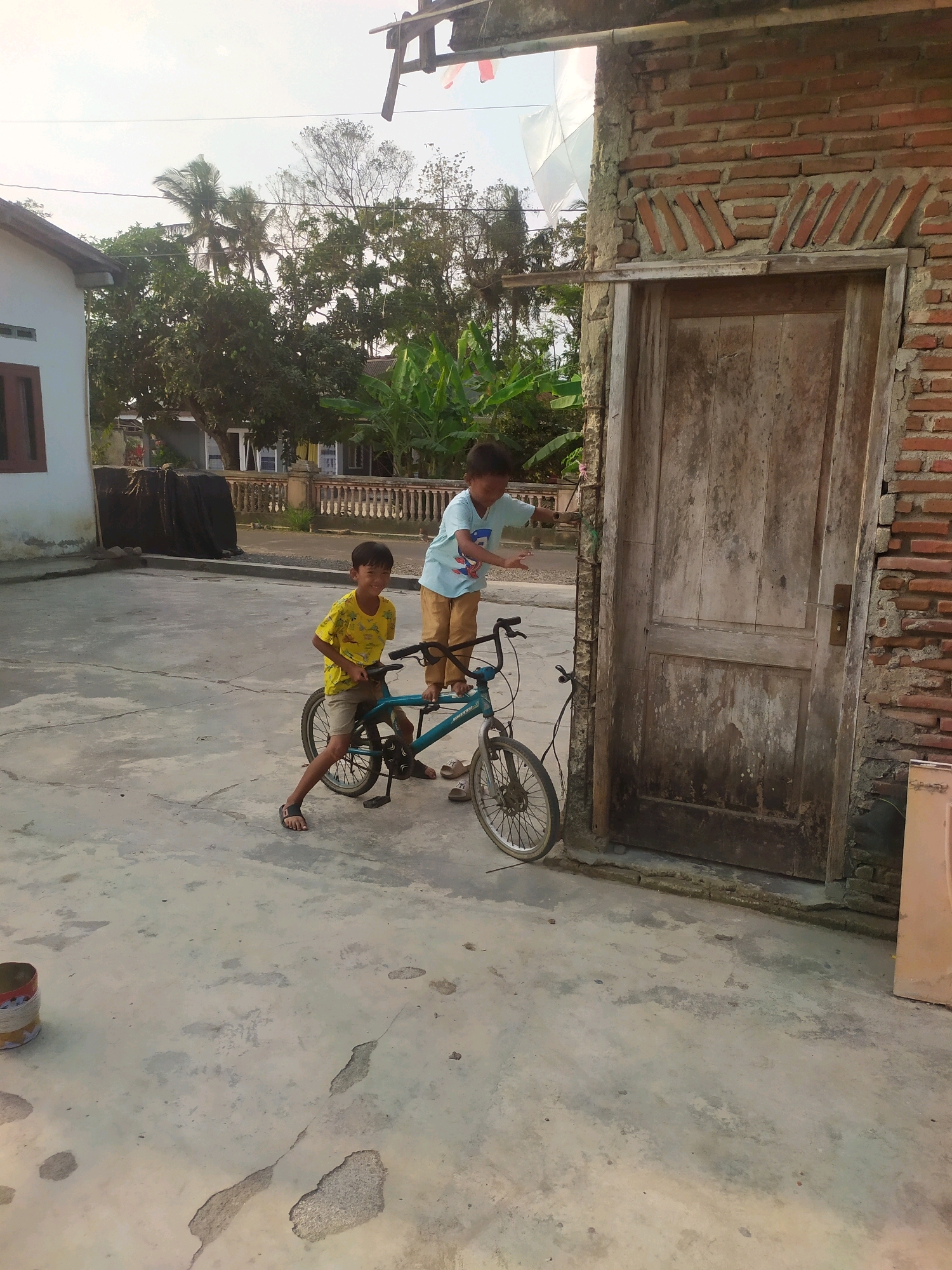 The image depicts an outdoor scene in Desa Penggalang Kecamatan Adipala, Indonesia, during daylight. In the foreground, two young boys are interacting. One boy, wearing a yellow t-shirt and shorts, is seated on a teal bicycle. The other boy, wearing a light blue t-shirt and brown pants, stands next to the bicycle, gesturing towards it. The setting appears to be a paved area with a weathered, wooden door set into a brick wall on the right side of the image. In the background, there is a white building with a brown window on the left, and a fence with decorative pillars. Lush green trees and palm trees are visible beyond the buildings, suggesting a tropical environment. The sky is bright and overcast. A metal can is visible on the ground in the lower left corner.