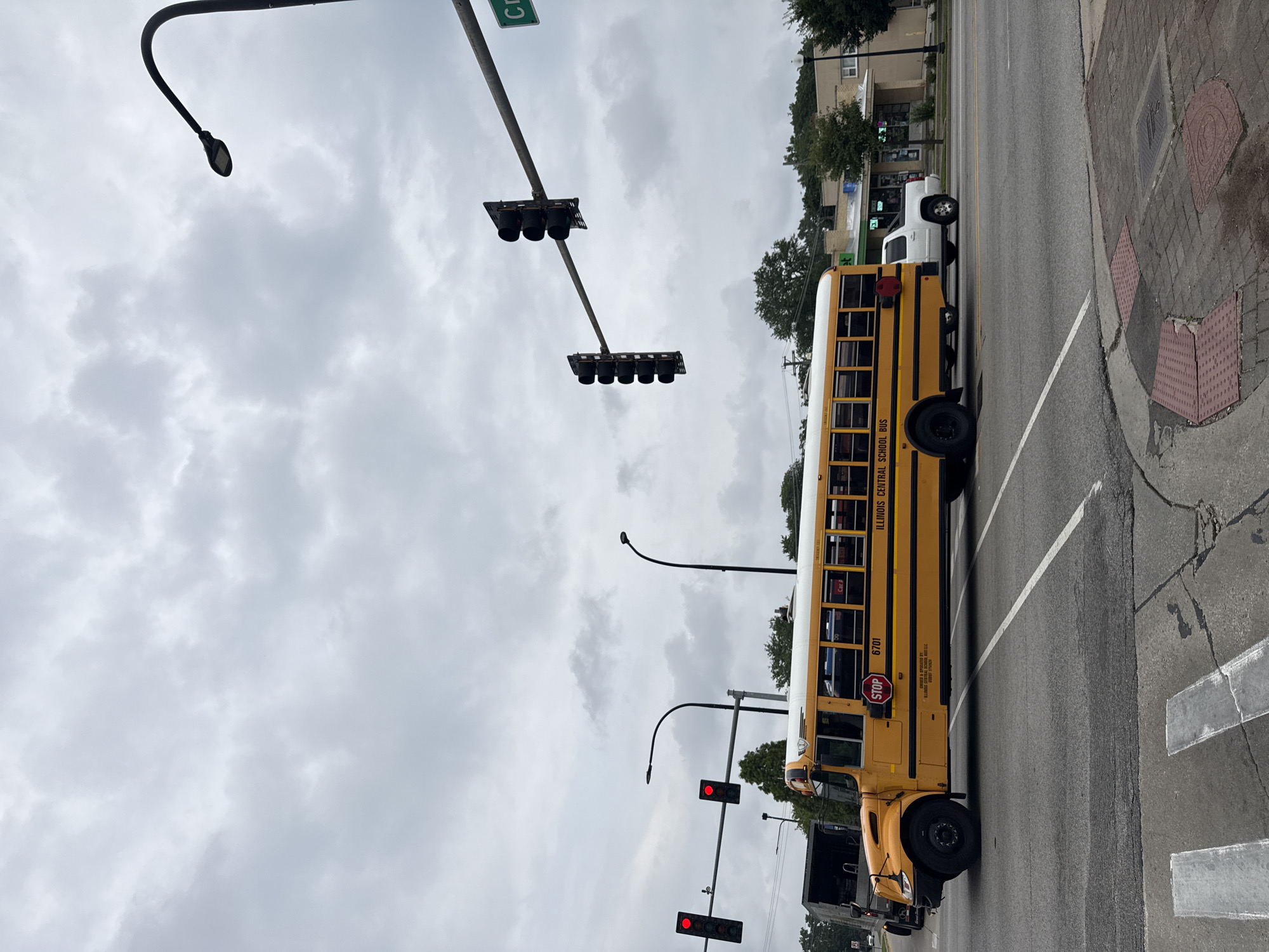 A yellow Illinois Central school bus with the number 6701 on its side is traveling on a four-lane road under an overcast sky. The bus is in the right lane, and a white pickup truck is following it. Traffic lights are visible overhead, with the lights for the bus's direction of travel appearing to be red. In the background, there are trees and buildings, suggesting an urban or suburban environment. A street sign visible in the distance reads "CR". The bus has its stop sign extended, indicating it may be stopped or about to stop.