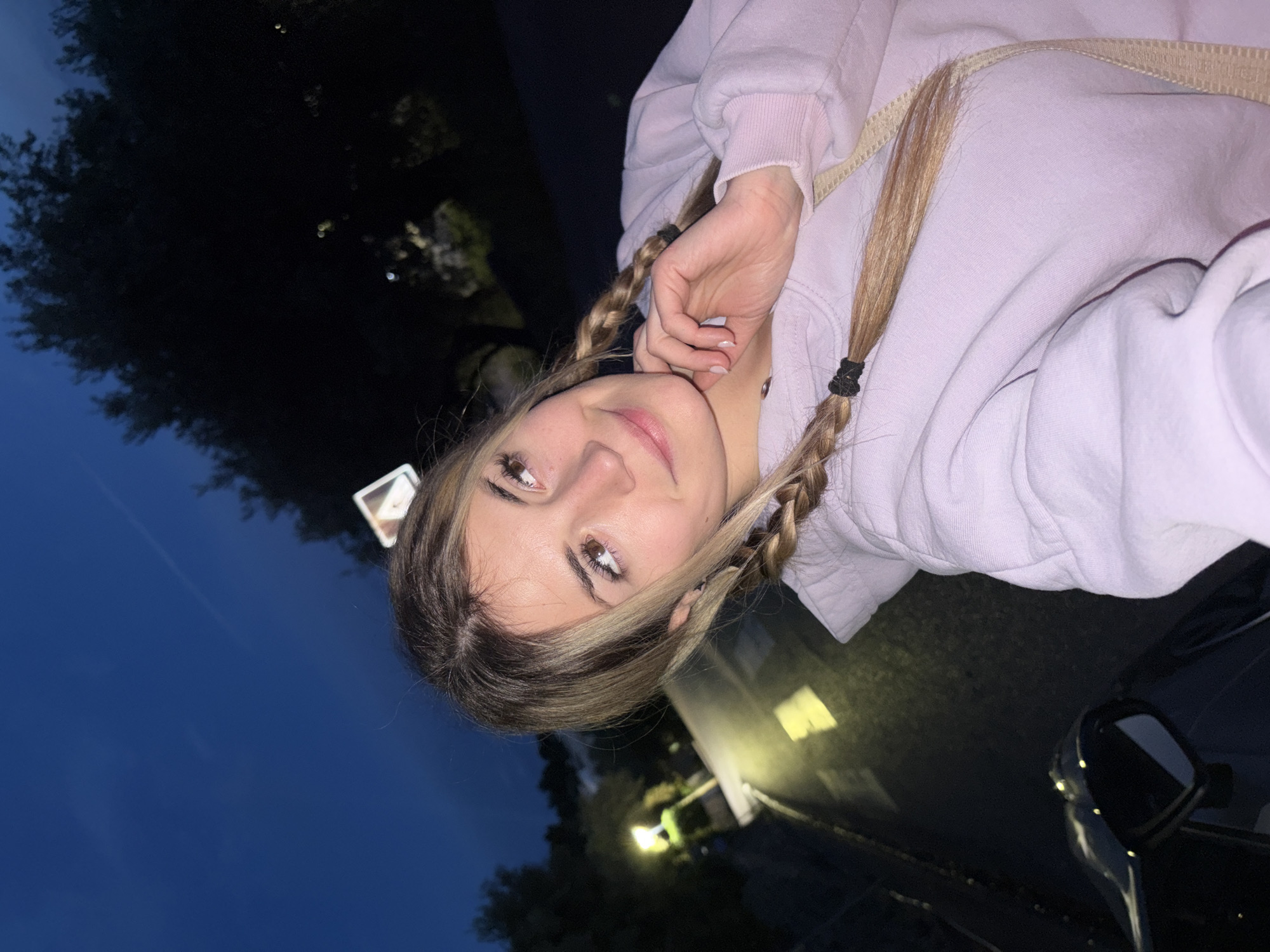 A young woman with brown eyes and blonde hair in braids is photographed at night. She wears a pale purple hoodie and appears to be in or near a car. The background shows trees, a blue night sky, and some structures that suggest an urban or suburban setting.