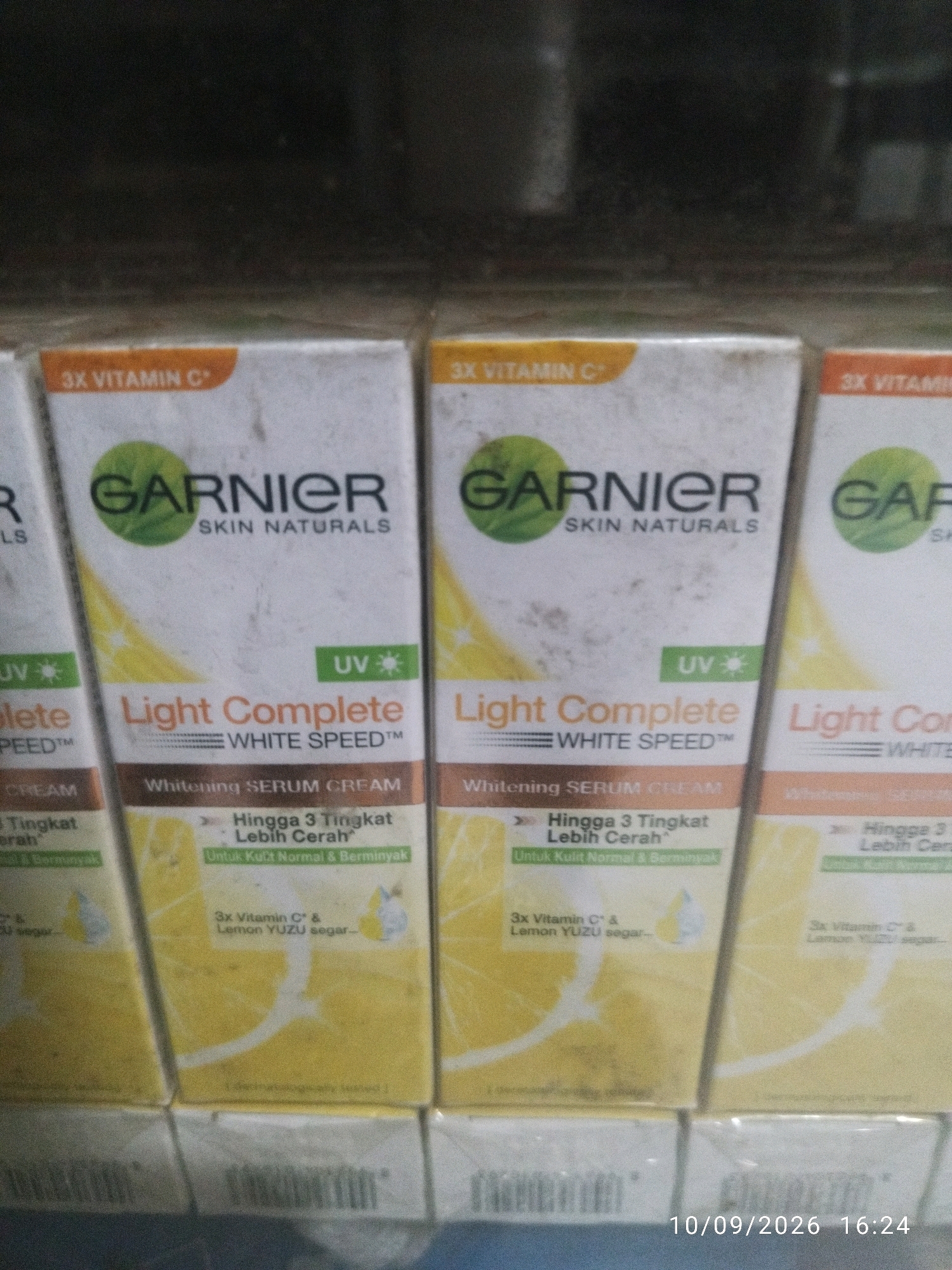 The image shows a close-up, eye-level view of multiple Garnier Skin Naturals Light Complete White Speed serum cream boxes arranged in a retail setting. The boxes are predominantly white with yellow accents, featuring the Garnier logo and text in Indonesian. The lighting appears to be artificial, casting a slight yellow hue on the products. In the bottom right corner, a timestamp of "10/09/2026 16:24" is visible, indicating the time and date the photo was taken. The background is dark and out of focus, suggesting a shelf or display unit. There are no people or animals visible in the image. The scene depicts products on display for sale, likely in a store or market.