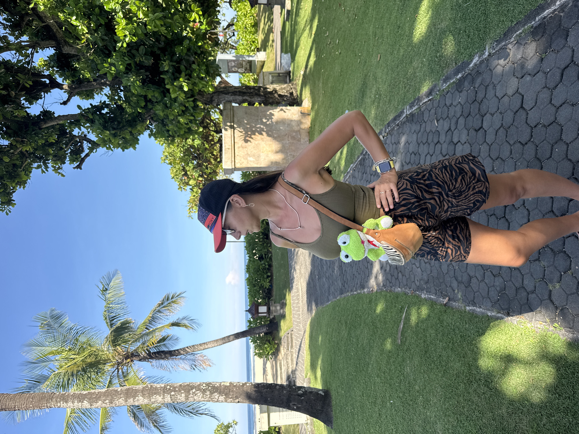 A person, presumably female, stands on a hexagonal-paved path with adjacent green grass in Kuta Selatan, Indonesia. Palm trees and a blue sky with faint clouds are visible in the background, along with a distant beach and ocean. The person is wearing a baseball cap, sunglasses, a light green tank top, and a brown and black patterned skirt. A brown purse with a plush green frog attached hangs across their torso. The scene suggests a tropical or coastal environment within a park or landscaped area. A building or monument can be seen behind the person.