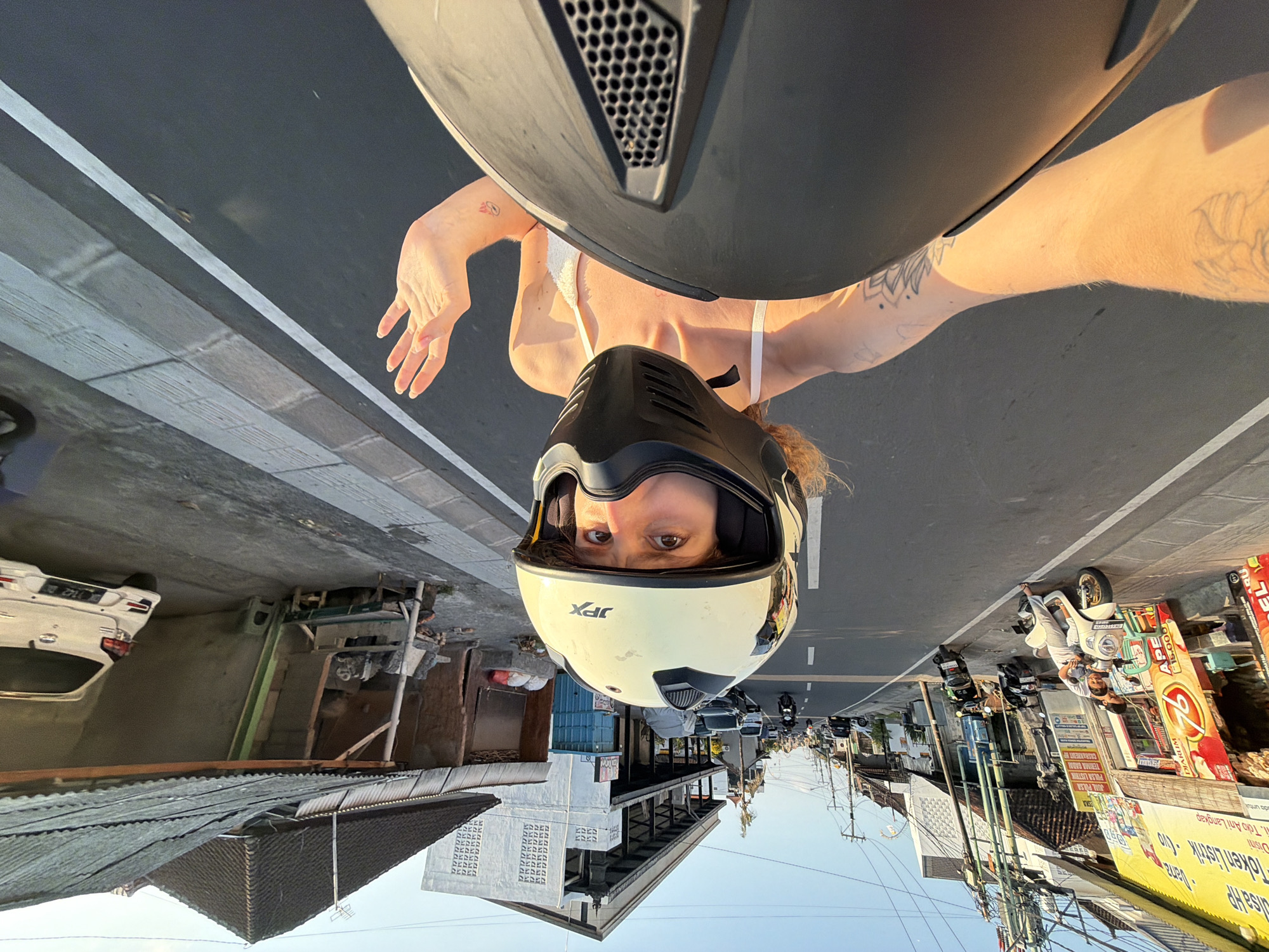 This is an upside-down, eye-level selfie of a person wearing a white bikini top and a black and yellow JPX motorcycle helmet. The subject's face is visible through the helmet's opening, with their eyes looking directly at the camera. Their arms are extended, with one hand visible in the foreground and tattoos on their arms.
The scene is a street in Mengwi, Indonesia, with a paved road and a sidewalk lined with buildings. The time of day appears to be late afternoon or early evening, as indicated by the warm, golden sunlight casting long shadows. The weather seems clear and sunny.
Various objects are visible, including parked motorcycles, cars, and a white car with an Indonesian license plate. Several shops and stalls line the street, with colorful signs and awnings. Some of the visible text on the signs includes "Token Listrik", "PULSA LISTRIK", and brand names. The overall impression is a bustling street scene in a tropical location. The perspective of the photo, being upside down, gives it a unique and disorienting feel.