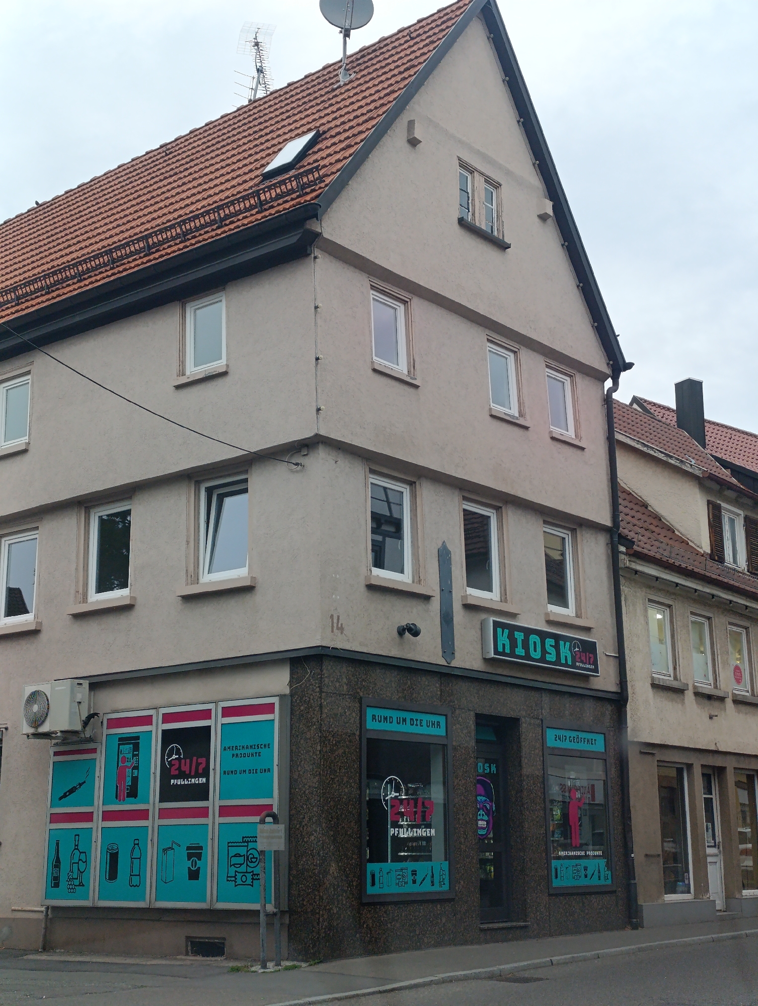 The image shows a corner view of a building in Pfullingen, Germany, with a KIOSK sign prominently displayed. The building has a beige facade and a red tiled roof. Several windows are visible on different levels of the building. The KIOSK appears to be a shop or convenience store, as indicated by the signage and the products displayed in the windows. One window showcases a selection of American products, with text stating "AMERIKANISCHE PRODUKTE RUND UM DIE UHR" (American products around the clock) and "24/7 GEÖFFNET" (24/7 open). Another window features graphics of various items like a vape, a vending machine, drinks, and snacks. A large, colorful graphic of a gorilla's head is visible in a doorway. The street in front of the building is paved, and there is a sidewalk. An air conditioning unit is mounted on the exterior wall of the building. The overall scene suggests a typical urban or suburban commercial area.