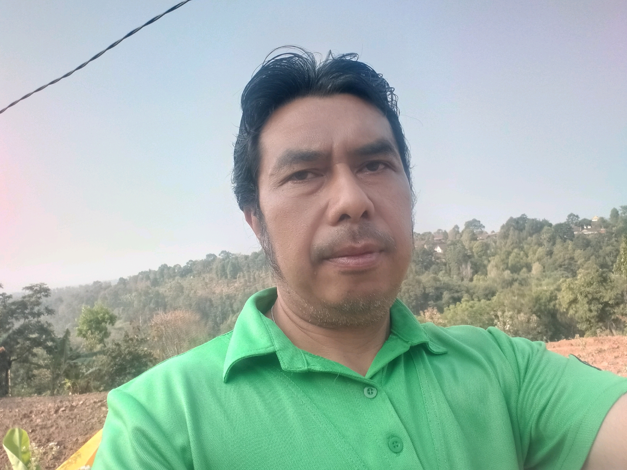 In a selfie taken outdoors, a man of Asian descent with dark hair and a light complexion is captured from the chest up. He is wearing a bright green collared shirt with two buttons visible. His facial expression is neutral, and his gaze is directed towards the camera. A thin wire stretches diagonally across the upper portion of the frame. The background features a hilly landscape covered with lush green trees, indicating a rural or natural setting. The sky is a pale blue, suggesting it is either early morning or late afternoon. The ground in the foreground appears to be dirt and possibly dry vegetation. The man's location is identified as Sawahan, Indonesia, a subdistrict in the Nganjuk Regency of East Java.