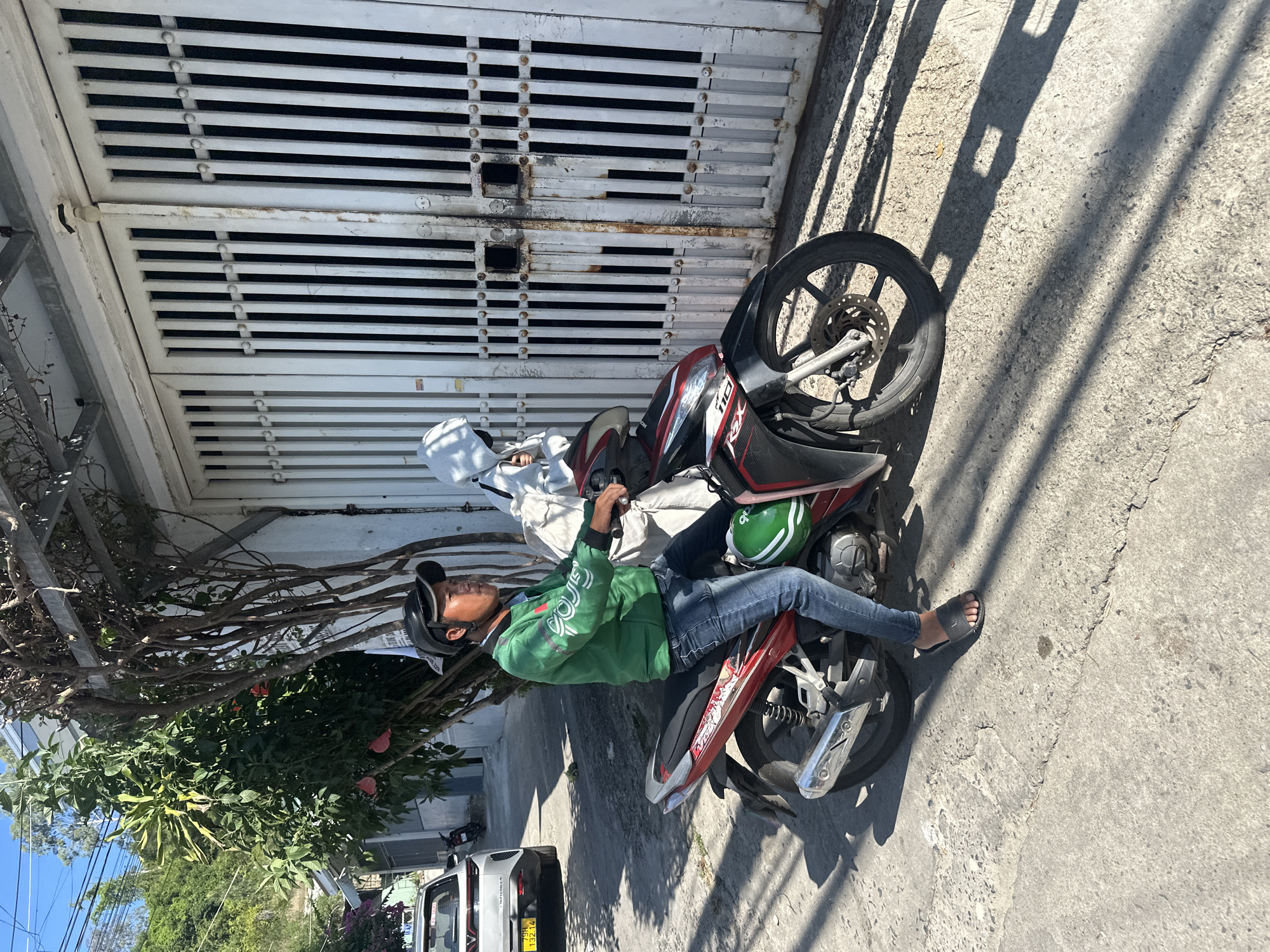 In Bac Nha Trang Ward, Vietnam, a man wearing a green jacket, blue jeans, and a black cap rides a red and black motorcycle. A passenger, dressed in a white hooded jacket and mask, sits behind him. They are on a paved street with a white metal gate and a large tree with green leaves and red flowers visible. A white car with a yellow license plate is parked on the left side of the street, and other buildings and trees are in the background. The sun casts shadows on the ground.