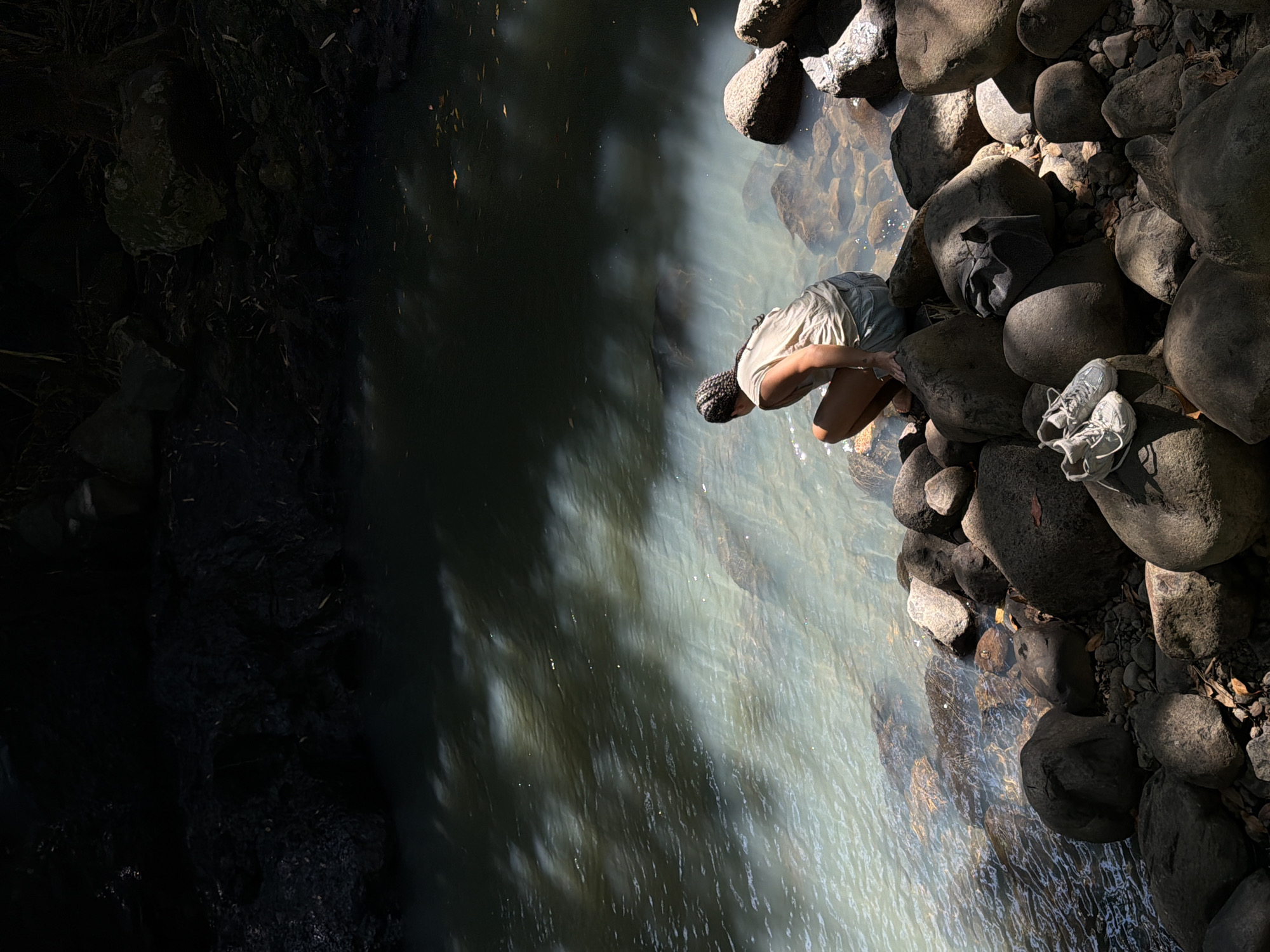 The image depicts a person squatting on large rocks at the edge of a body of water, likely a river, in Selemadeg, Indonesia. The person, wearing a light-colored tank top and denim shorts, is facing away from the camera and towards the water. Their hair is braided and they are wearing a patterned head wrap. Sunlight filters through trees, casting dappled shadows on the water and surrounding rocks. In the foreground, a pair of white athletic shoes and a dark-colored hat rest on the rocks. The water appears murky and reflects the surrounding foliage. The environment suggests a natural, possibly rural, setting with dense vegetation and rocky terrain.