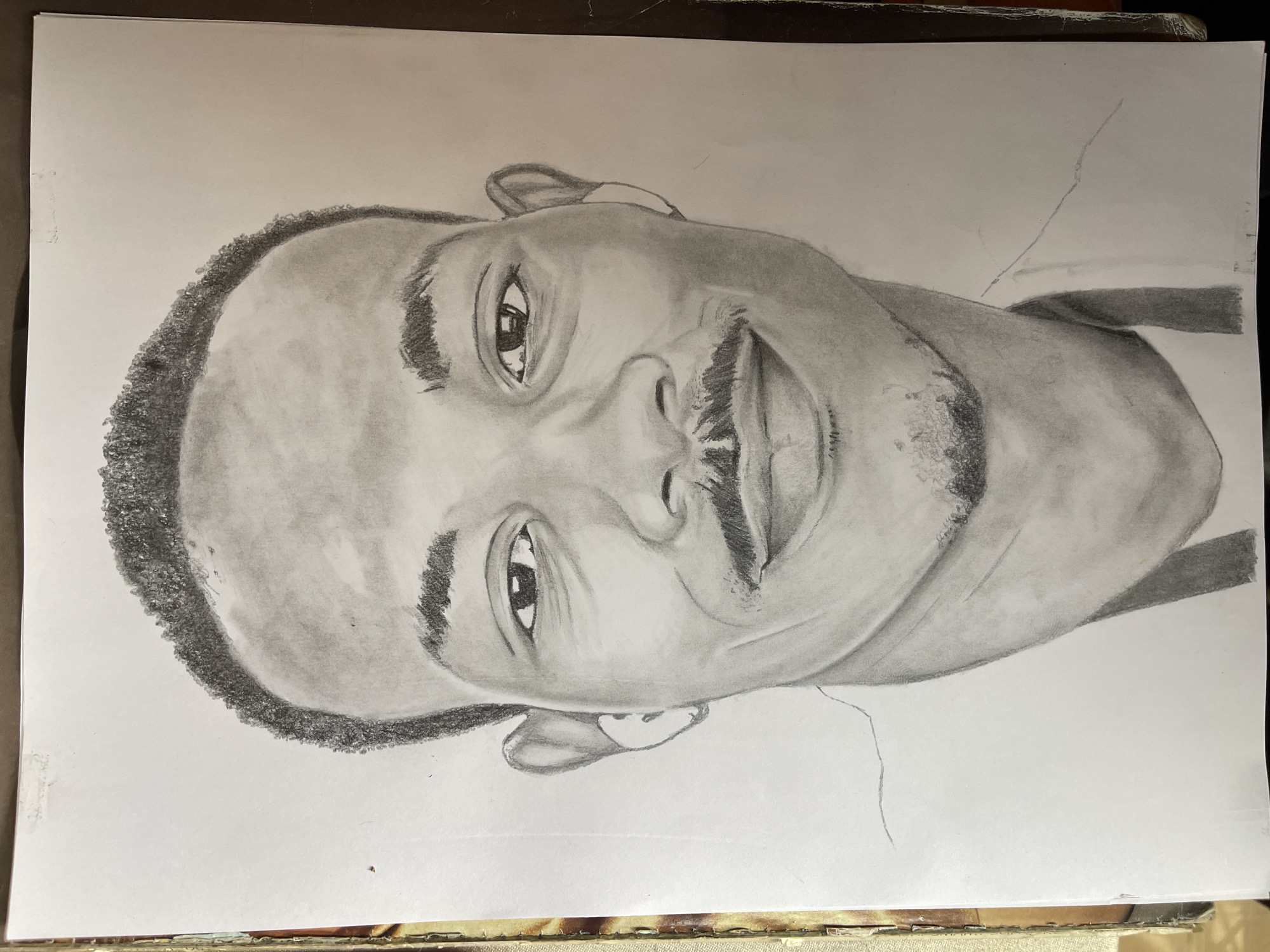 A portrait of a man with a closely cropped, textured hairline is drawn in graphite on white paper. He is depicted from the chest up, looking at the viewer with a smile. The drawing shows shading and detail in his eyes, nose, mouth, and ears. He also has short facial hair. The background is plain white paper. There is no evidence to suggest the location is Jos, Nigeria.