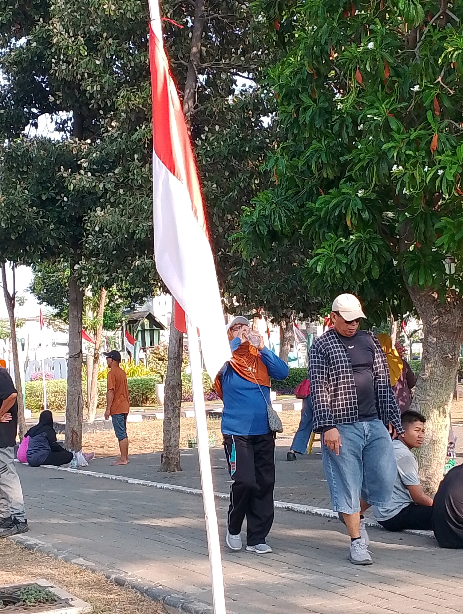 An outdoor scene is depicted, likely in Probolinggo, Indonesia, featuring a prominent Indonesian flag at the forefront. Several individuals are present. To the left, a person wearing a black shirt and grey pants stands with hands on hips. Next to them, a woman in a dark blue hijab and matching pants sits on the ground with a pink bag. Further back, a man in an orange t-shirt and blue shorts stands barefoot, facing away from the camera. In the mid-ground, a woman wearing an orange hijab and blue long-sleeved shirt drinks from a water bottle. To her right, a man in a checkered long-sleeved shirt, black t-shirt, and denim shorts walks towards the right, wearing a beige cap and white sneakers. Behind him, a person in a yellow hijab is partially visible, and a young man in a grey t-shirt and black shorts sits on the ground looking to his right. The environment includes trees with lush green foliage, a paved walkway, and grassy areas. A small building with a green roof is visible in the background.