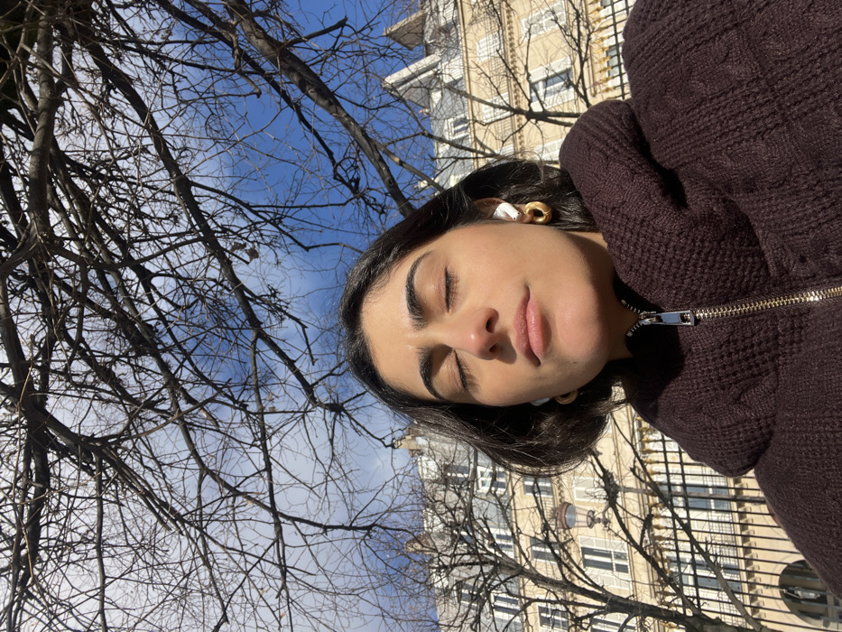 A medium close-up, low-angle shot captures an individual outdoors in Paris, France. The subject, a person with dark hair and a medium complexion, is positioned centrally in the lower frame, wearing a dark brown knitted jacket with a visible gold zipper. Their eyes are closed, and their face is angled slightly upward, illuminated by direct sunlight. White wireless earbuds are present in both ears, alongside gold-colored hoop earrings. The upper two-thirds of the image is dominated by a dense network of bare, dark tree branches silhouetted against a bright blue sky with scattered thin white clouds. In the mid-ground, behind the trees, are multi-story Haussmannian buildings characteristic of Parisian architecture, featuring light-colored stone facades, numerous windows, and decorative elements. A black metal fence and a vintage-style streetlamp are visible in the background, beneath the trees and in front of the buildings.