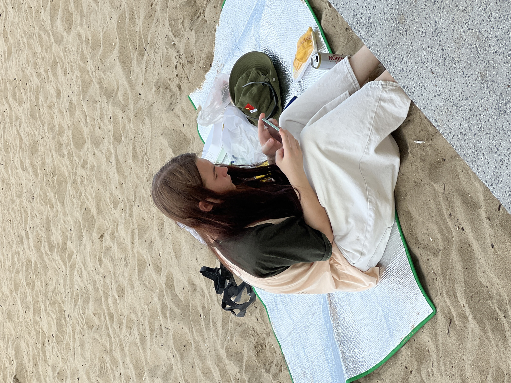 A young person with long brown hair is sitting on a silver and green picnic blanket on a sandy beach. They are wearing a light peach and dark green t-shirt and loose cream-colored pants. The person is looking down at a smartphone they are holding in their hands. To their left, a pair of black sandals are lying on the sand. On the picnic blanket, there is a green hat with a red star on it, a plastic bag, a can of Saigon beer, and a small rectangular container of yellow food. The background is a sandy beach with footprints. The scene appears to be in Nha Trang, Vietnam, as indicated by the Saigon beer can and the style of the hat.