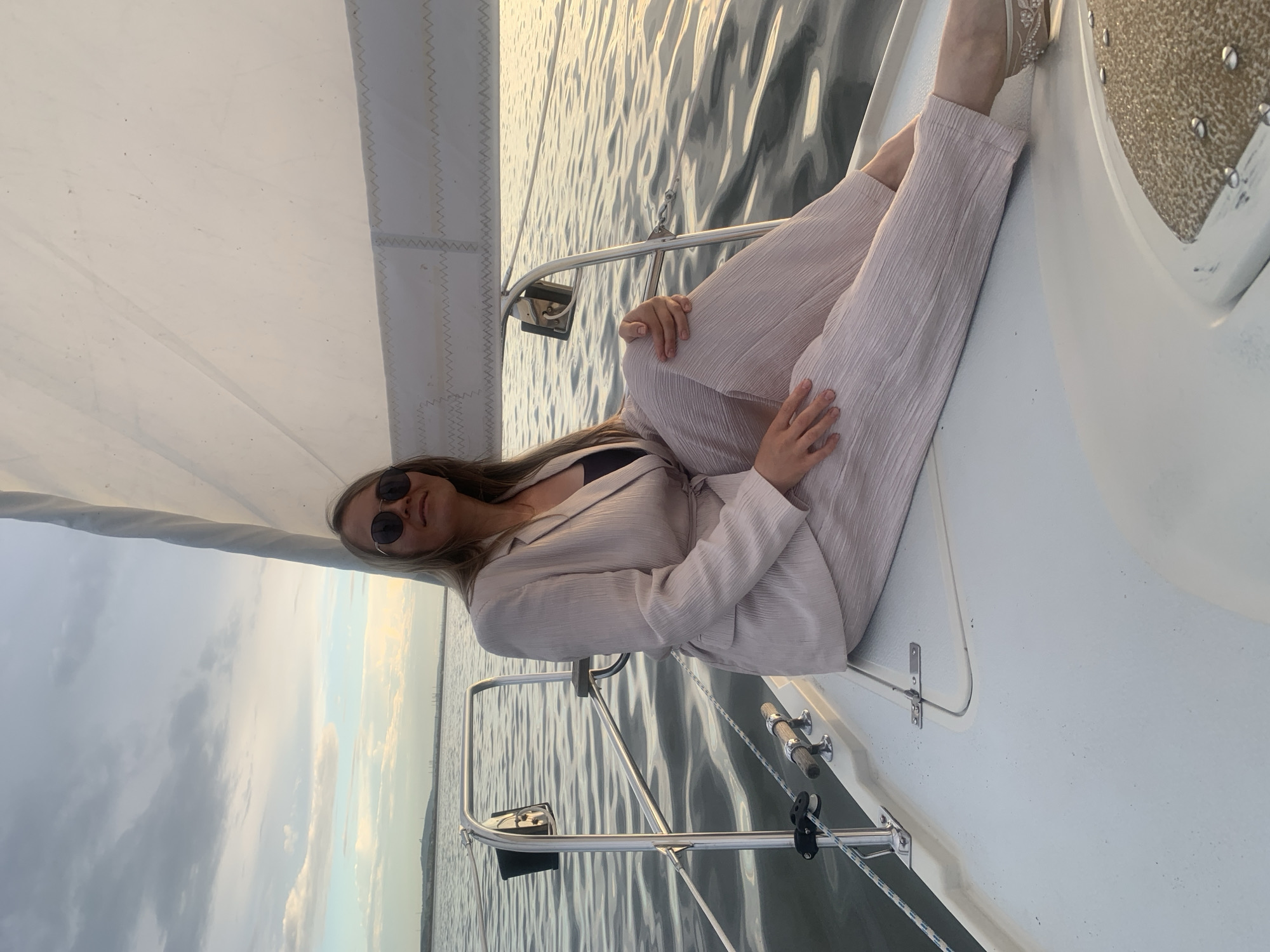 A woman wearing a light pink pantsuit and sunglasses sits on the bow of a sailboat. The boat is on the water, with a sail visible behind her. The sky is cloudy with hints of sunset colors. The location is the Kama River in Chaykovsky, Russia.