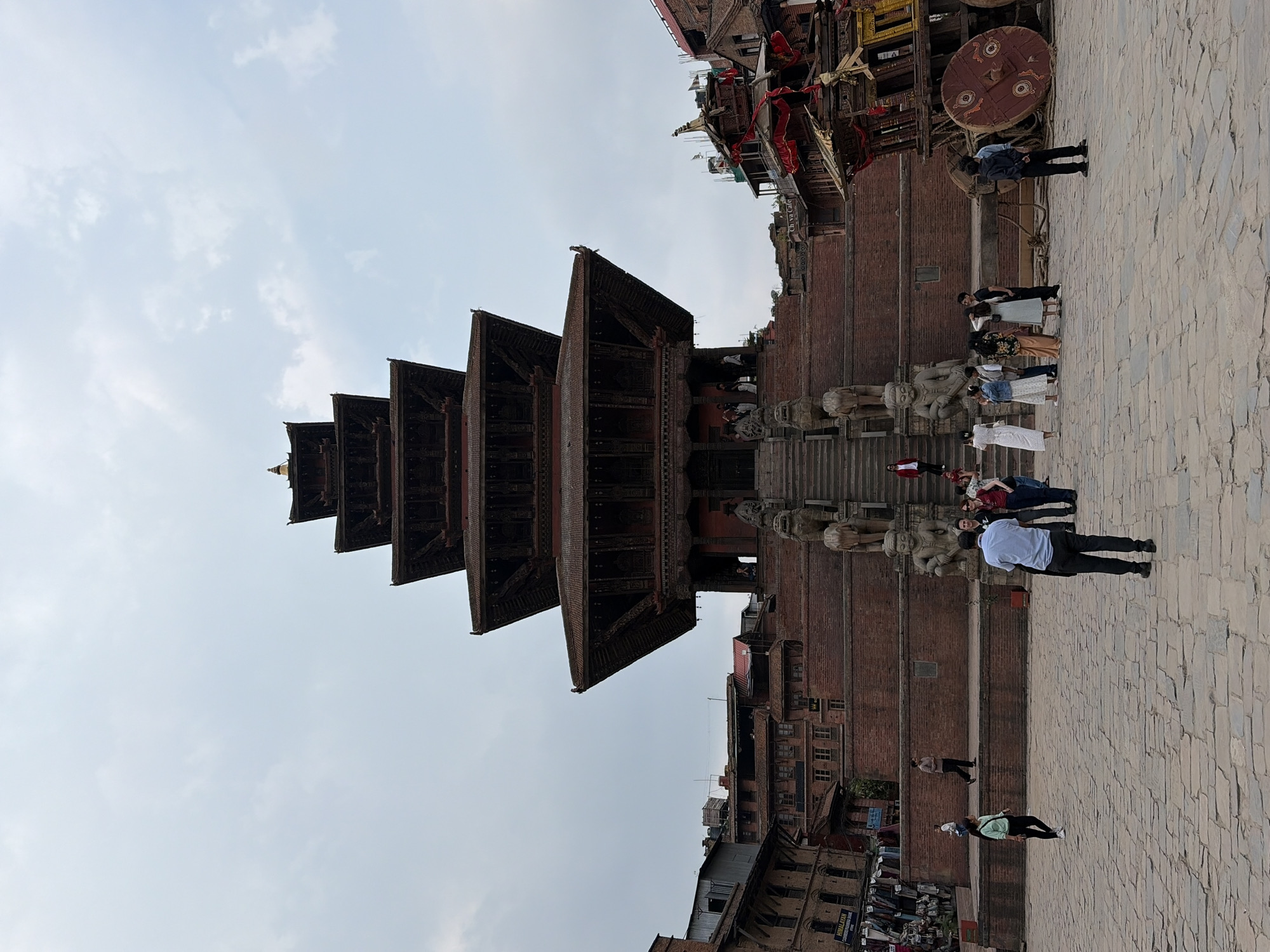 The sideways-oriented image captures a wide view of a multi-tiered pagoda temple dominating the left side of a bustling square, set against a soft, cloudy sky. The temple, constructed from dark wood and brick, features at least five prominent tiered roofs adorned with intricate carvings and topped with a golden finial. A grand stone staircase leads up to the temple's entrance, flanked by large, ornate stone statues, likely guardians or deities.
To the right of the temple, a large public square is paved with light-colored stones. Numerous people are present, indicating a lively atmosphere. Some individuals are gathered near the base of the temple's steps, while others are walking or standing across the plaza. In the mid-ground right, a man in a light blue shirt and dark pants stands with his back to the camera. Further into the background, traditional brick and wooden buildings line the square, housing various establishments. A large, ornate wooden wheel, possibly a ceremonial chariot wheel, is prominently displayed in the upper right portion of the image.
The scene takes place during the daytime under an overcast sky, casting a soft, even light. Visible text includes "Chiya Cafe" on a sign attached to a building, and "Himalayan Store" partially visible on another sign further down. The architecture and general setting are characteristic of Bhaktapur Durbar Square in Nepal.