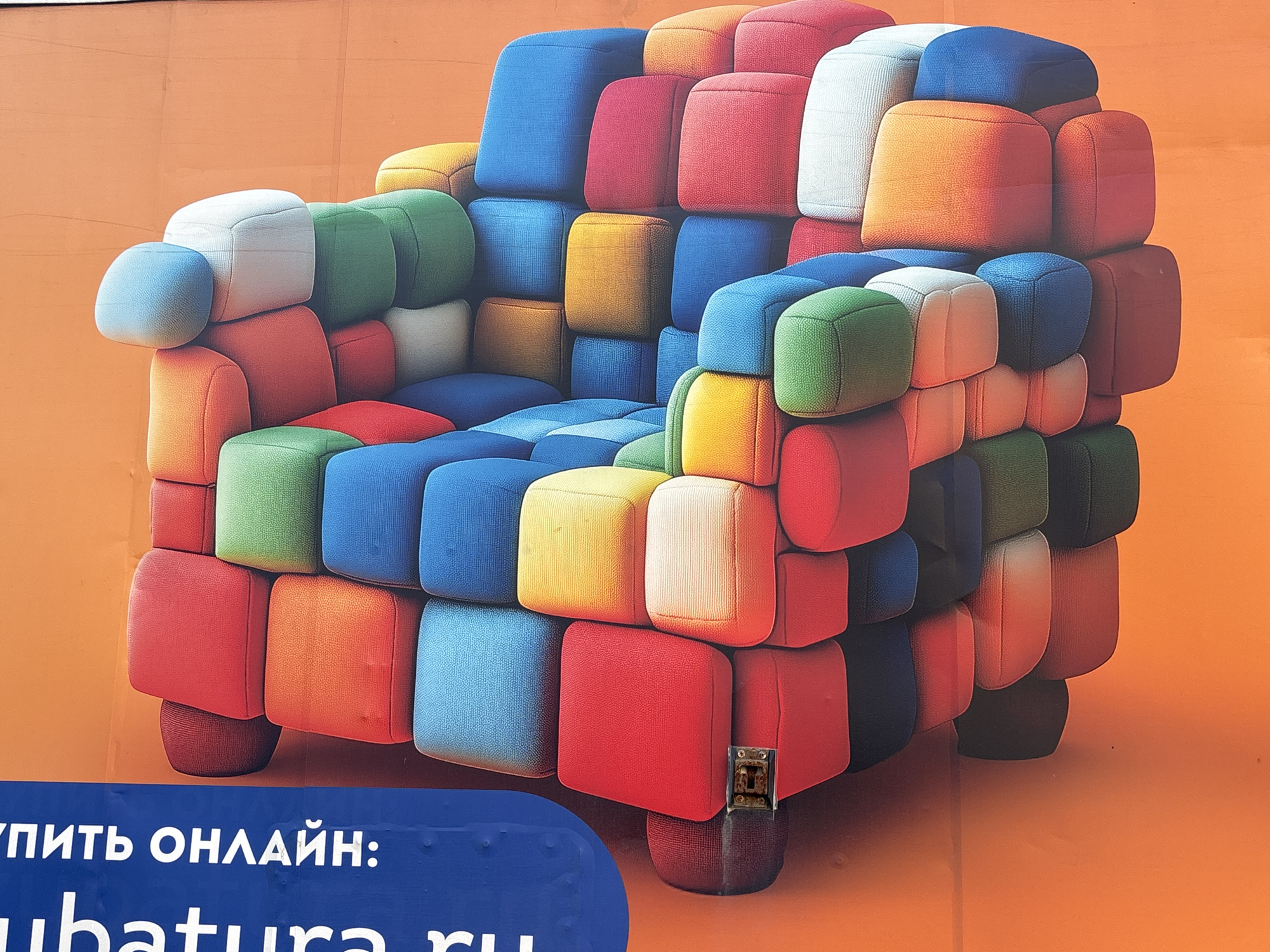 A photograph shows a vividly colored armchair seemingly constructed from multi-hued padded cubes of red, blue, green, orange, yellow, and white. The background is a solid orange color. The armchair appears to be a piece of advertising material. The phrase "КУПИТЬ ОНЛАЙН:" and part of the website address "ubatura.ru" are visible in the lower left corner on a blue background, indicating an online retail location. The context clues suggest this might be an advertisement in Saint Petersburg, Russia, for the chair.