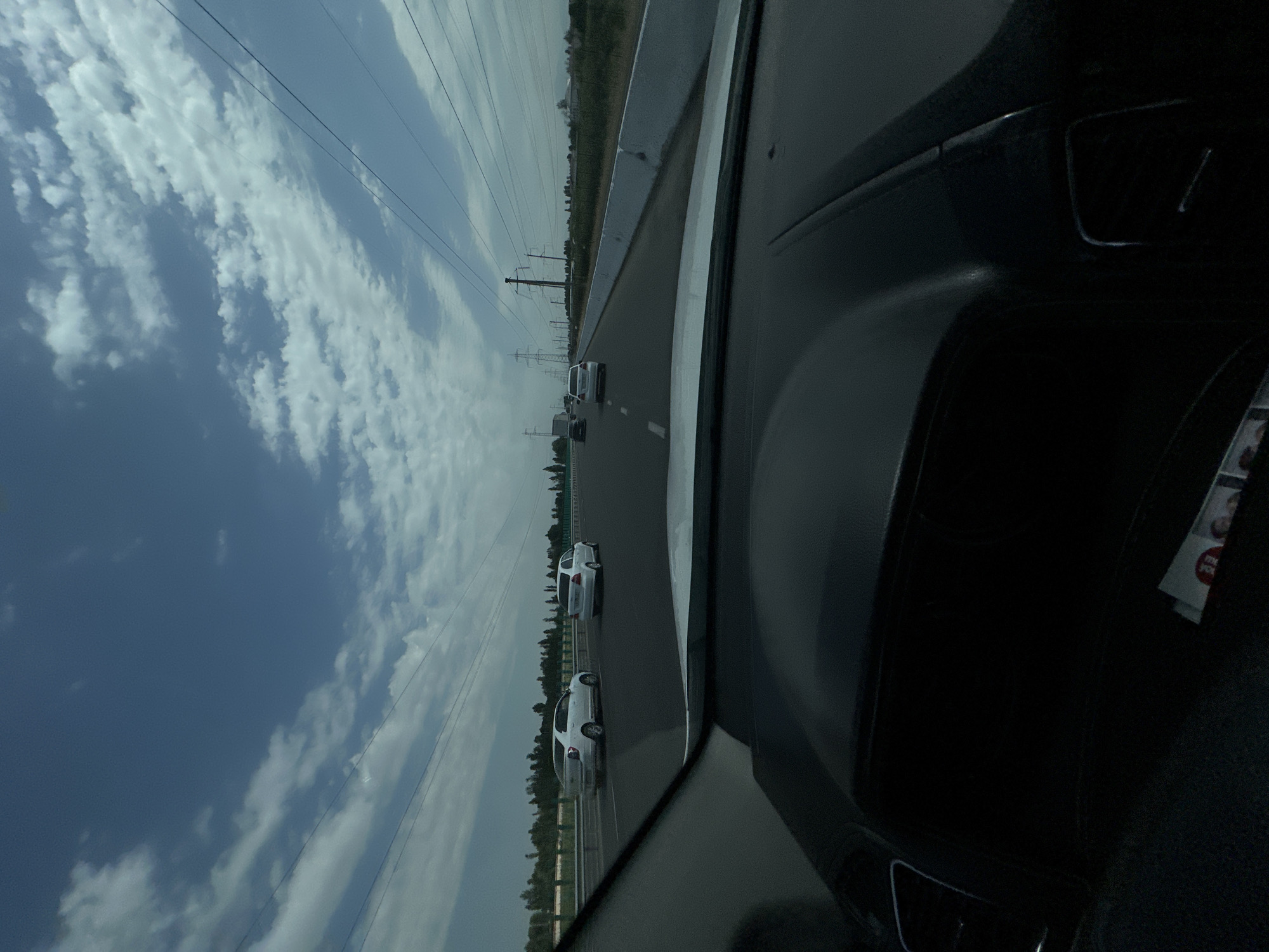 The image depicts a view from inside a vehicle traveling on a highway. The sky is a bright blue with scattered white clouds. There are several cars on the road ahead, all moving in the same direction. Power lines and utility poles are visible along the side of the road, as well as trees and fields in the distance. The dashboard of the vehicle is visible in the foreground, with some papers or magazines tucked into a compartment. The location is identified as Akbarak, Uzbekistan.
