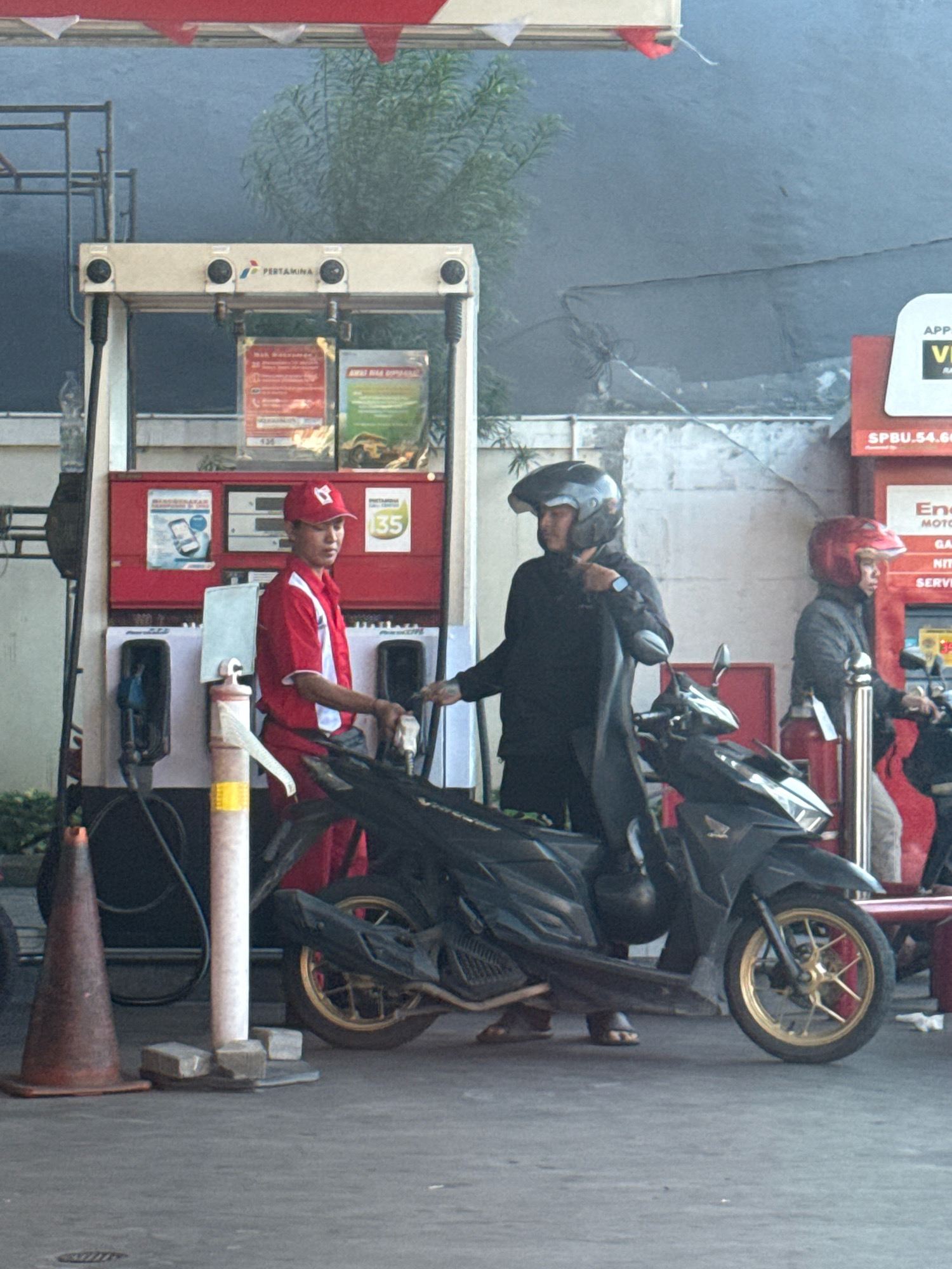 At a Pertamina gas station in Surabaya, Indonesia, a gas station attendant in a red uniform and cap is dispensing fuel into a motorcycle. The motorcycle is dark colored and has a rider wearing a black jacket and helmet. Another person in a red helmet is standing nearby, also on a motorcycle. The gas station has red and white dispensers and signage. The ground is paved.