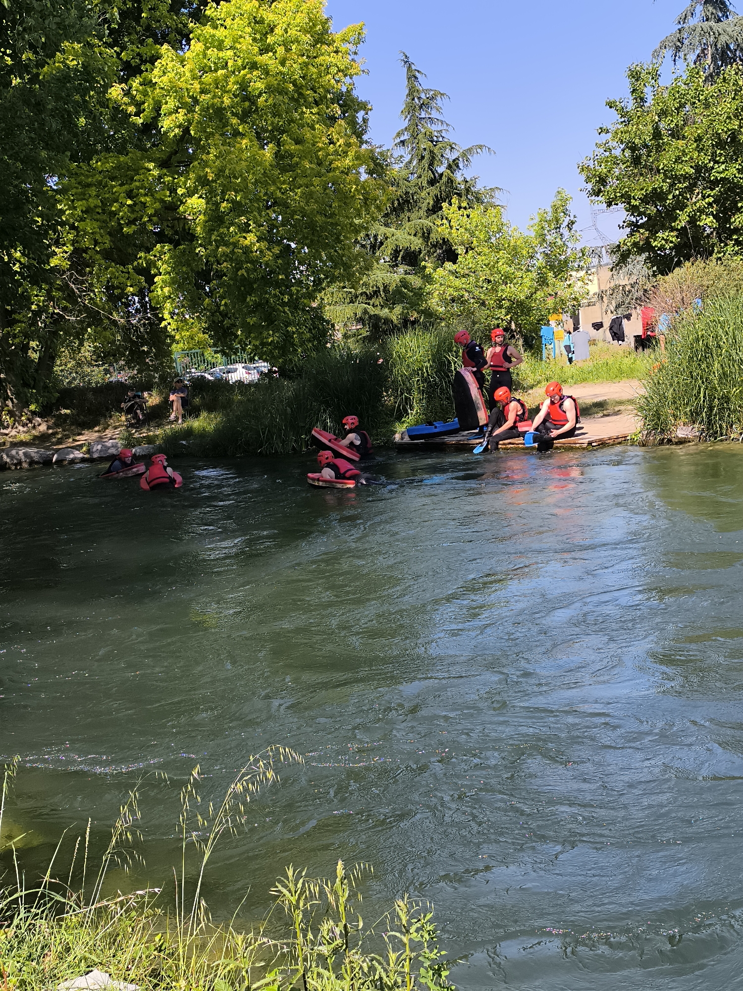 The scene captures a group of people engaged in river activities in Saint-Pierre-de-Bœuf, France. Several individuals wearing red helmets and life jackets are in the water, some on floating boards. Others, also wearing red helmets and life jackets, are near the riverbank, possibly preparing for or concluding their water activities. Tall green trees and vegetation surround the river, with a building visible in the background. Some bystanders can be seen on the banks. The water is flowing and somewhat murky. Clothes are hanging and drying in the far background.
