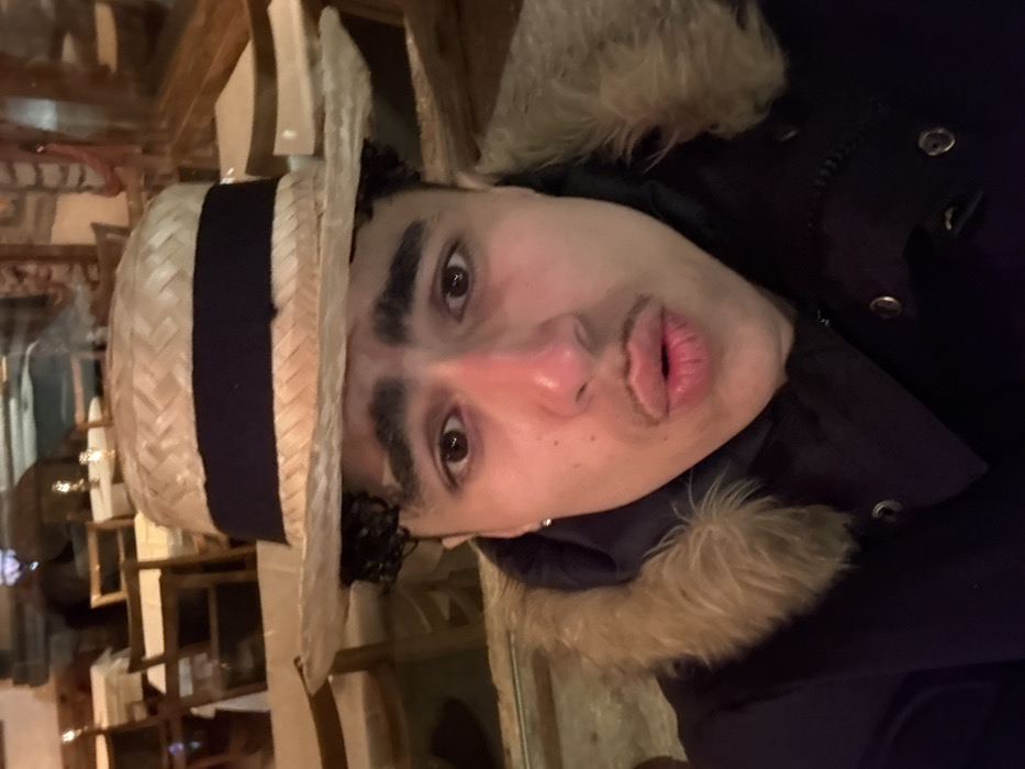 An individual is depicted in a close-up, chest-up frame in the city of La Clusaz, France. The person wears a straw boater hat with a dark band, a dark winter coat featuring a prominent light brown fur-lined hood, and a dark hoodie beneath. Visible features include dark curly hair, a short mustache above pursed lips forming an 'O' shape, wide-open eyes, and a silver stud earring in the left ear. The background consists of reflections showing an indoor environment, specifically what appears to be a restaurant or cafe interior with wooden chairs, tables covered with white tablecloths, and small illuminated objects, possibly candles or lamps.