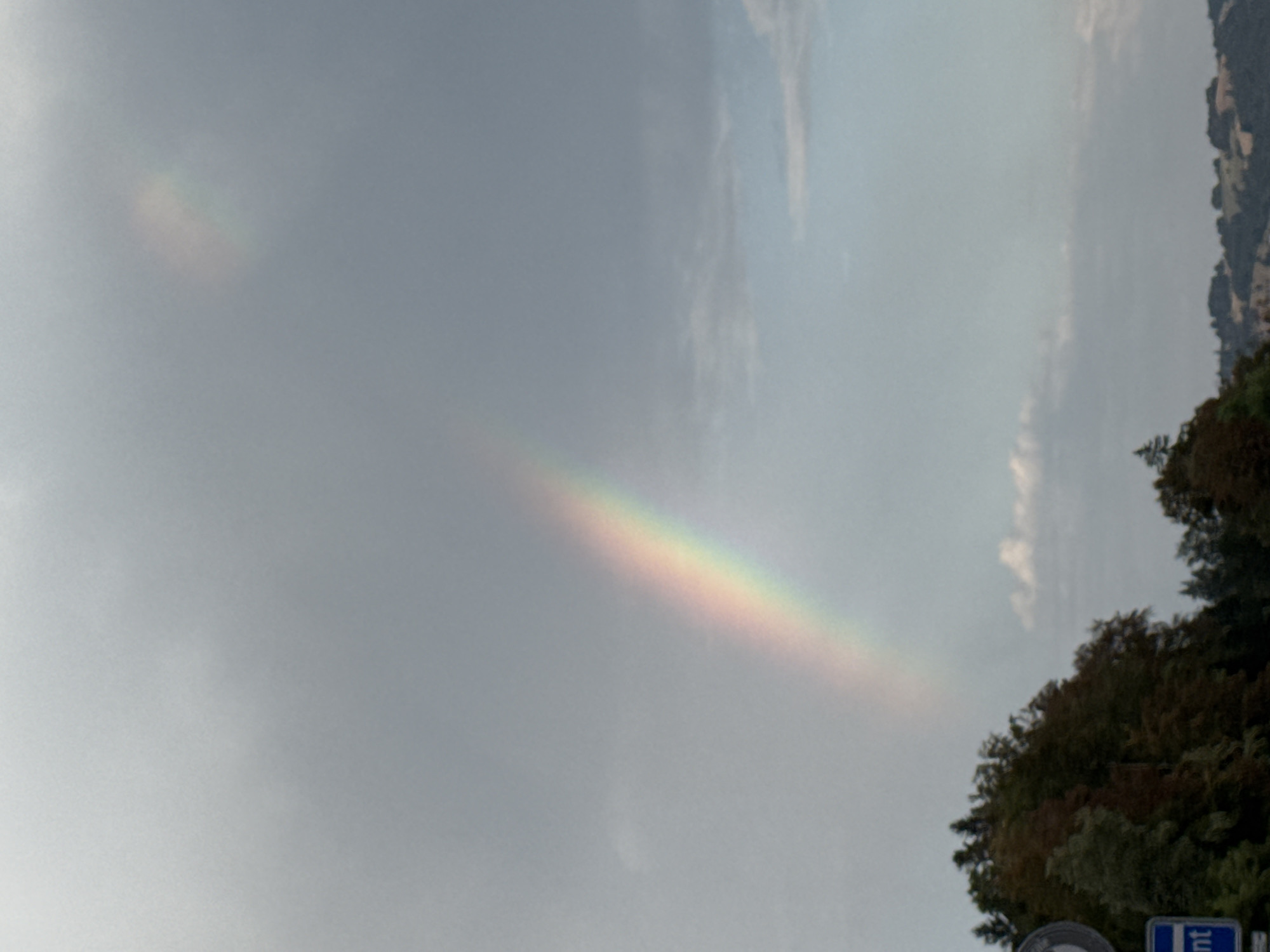 In the city of Bellelay, Switzerland, a faint rainbow arches across a cloudy, overcast sky. The rainbow is positioned towards the right of the image and appears to be a secondary rainbow due to its weaker intensity and reversed color order. The lower part of the image shows a treeline with a mix of evergreen and deciduous trees, some exhibiting autumn colors. A portion of a road sign is visible in the lower left corner, displaying a blue rectangular sign with white text that reads "nt". In the distance, rolling hills can be seen under the hazy sky. The overall scene suggests an outdoor, possibly rural setting in Bellelay during a time of intermittent rain and sunshine.