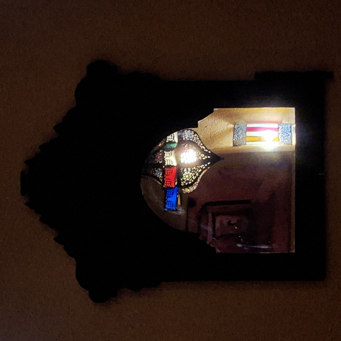 A dark, oddly-shaped frame, likely a mirror, hangs on a tan wall. Inside the frame, a scene reflects: a stained-glass lamp with red and blue panels sits within a room. The background features a window with colorful panes and a glimpse of furniture, giving depth to the reflection. The frame's irregular edges and color contrast draw attention to the reflective scene.