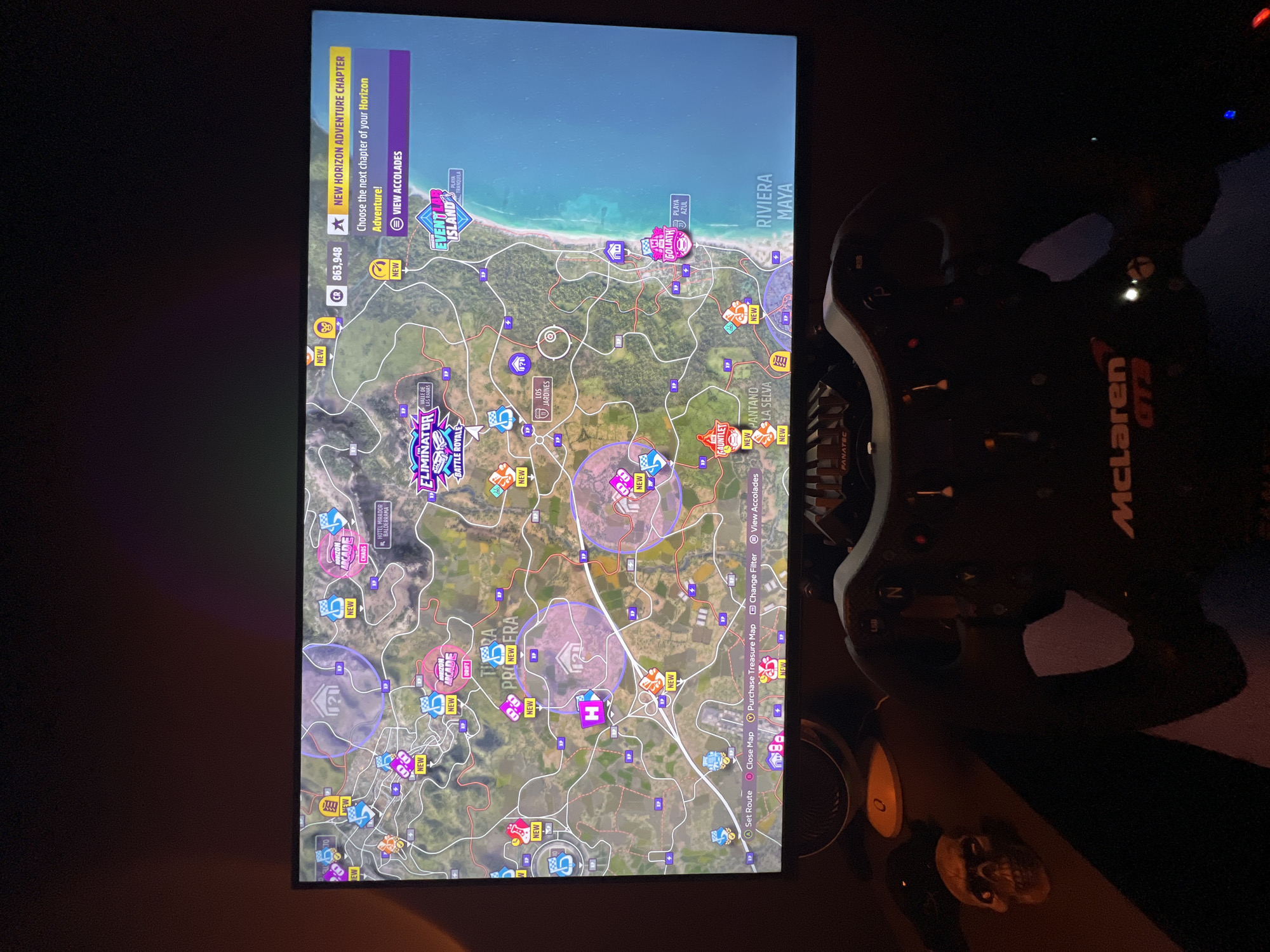 The image displays a gaming setup in a dimly lit room, likely at night. A vertically oriented screen shows a detailed video game map, presumably from Forza Horizon, featuring a vibrant, diverse landscape with coastal areas, roads, mountains, and forests. The in-game location is identified as "RIVIERA MAYA," with specific points like "PLAYA AZUL," "PANTANO LA SELVA," "LOS JARDINES," and "HOTEL MIRADOR BALDERRAMA," indicating a Mexico-inspired setting.
The map is densely populated with various event markers and points of interest, many highlighted with a "NEW" tag. Visible event types include "ELIMINATOR BATTLE ROYALE," "EVENT LAB ISLAND," and a large "GOLIATH" race. The user interface on the top left of the screen shows "CR 863,948" (in-game currency), a prompt for a "NEW HORIZON ADVENTURE CHAPTER," and an option to "VIEW ACCOLADES." Map controls at the bottom right offer functions such as "Set Route," "Close Map," "Purchase Treasure Map," "Change Filter," and "View Accolades."
To the right of the screen, a racing wheel peripheral is partially visible, distinctly branded with "FANATEC" and "McLaren GT3" on its rim. Other discernible objects include a dark speaker and a small, light-colored skull decoration. The overall activity depicted is video gaming. The physical location where the image was captured is Budva, Montenegro.